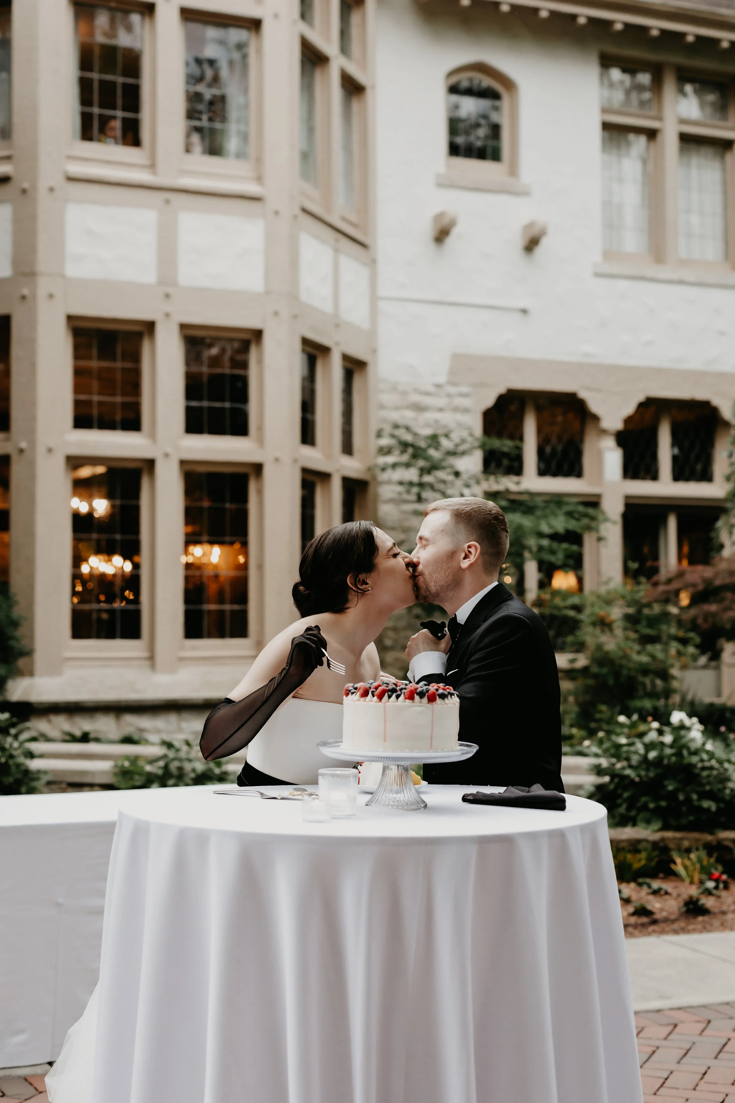 Bride and groom sharing a kiss beside their wedding cake in the courtyard of the Ohio Governor’s Residence, photographed by Julia Brown Photo.