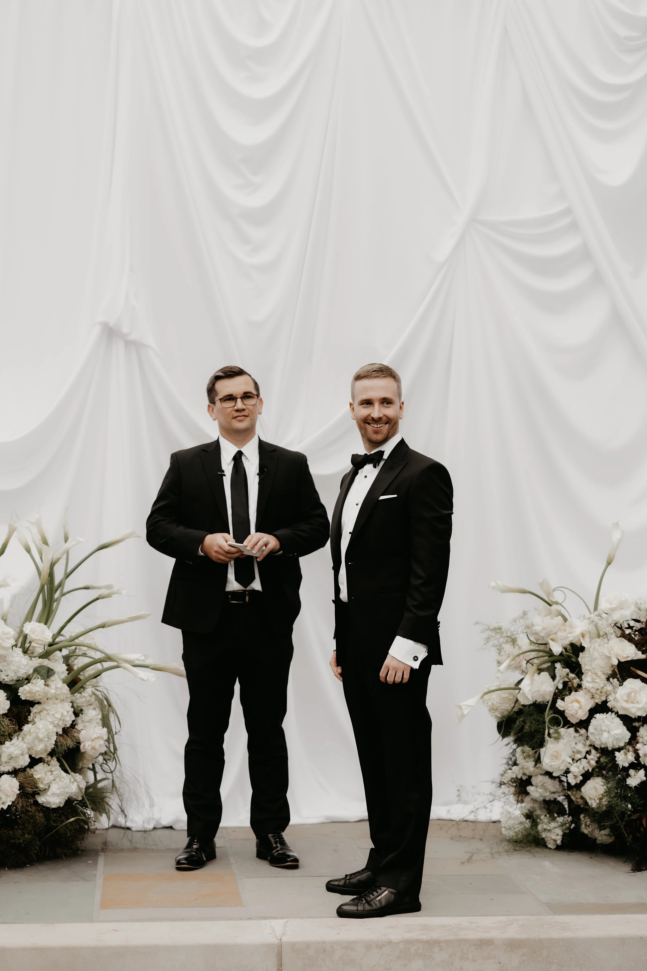 Groom and a wedding attendant standing before a white draped ceremony backdrop and organic floral installations, photographed by Julia Brown Photo.