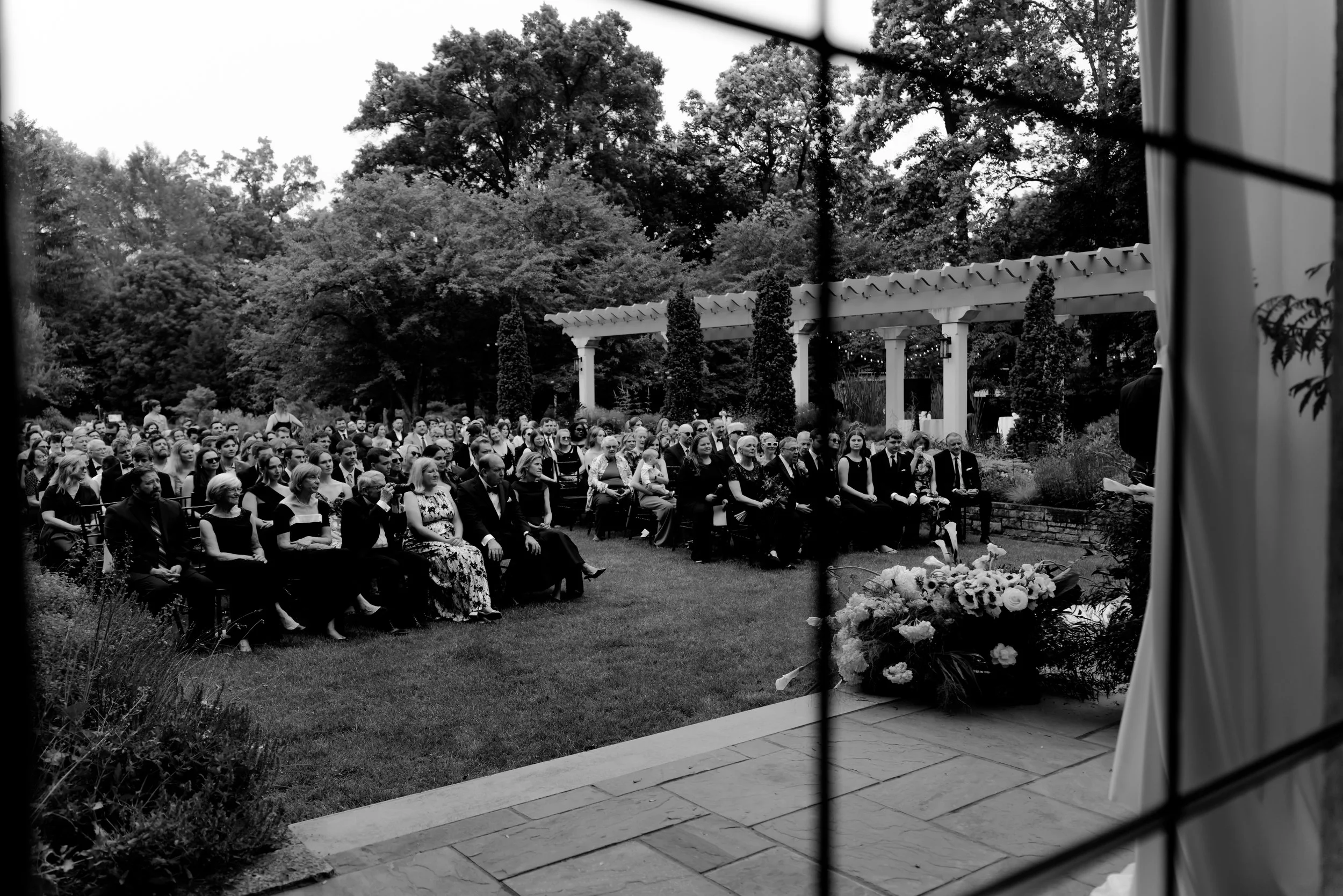 Black-and-white photograph of guests taken through a window watching the bride and groom’s outdoor wedding ceremony in the Heritage Garden, photographed by Julia Brown Photo.