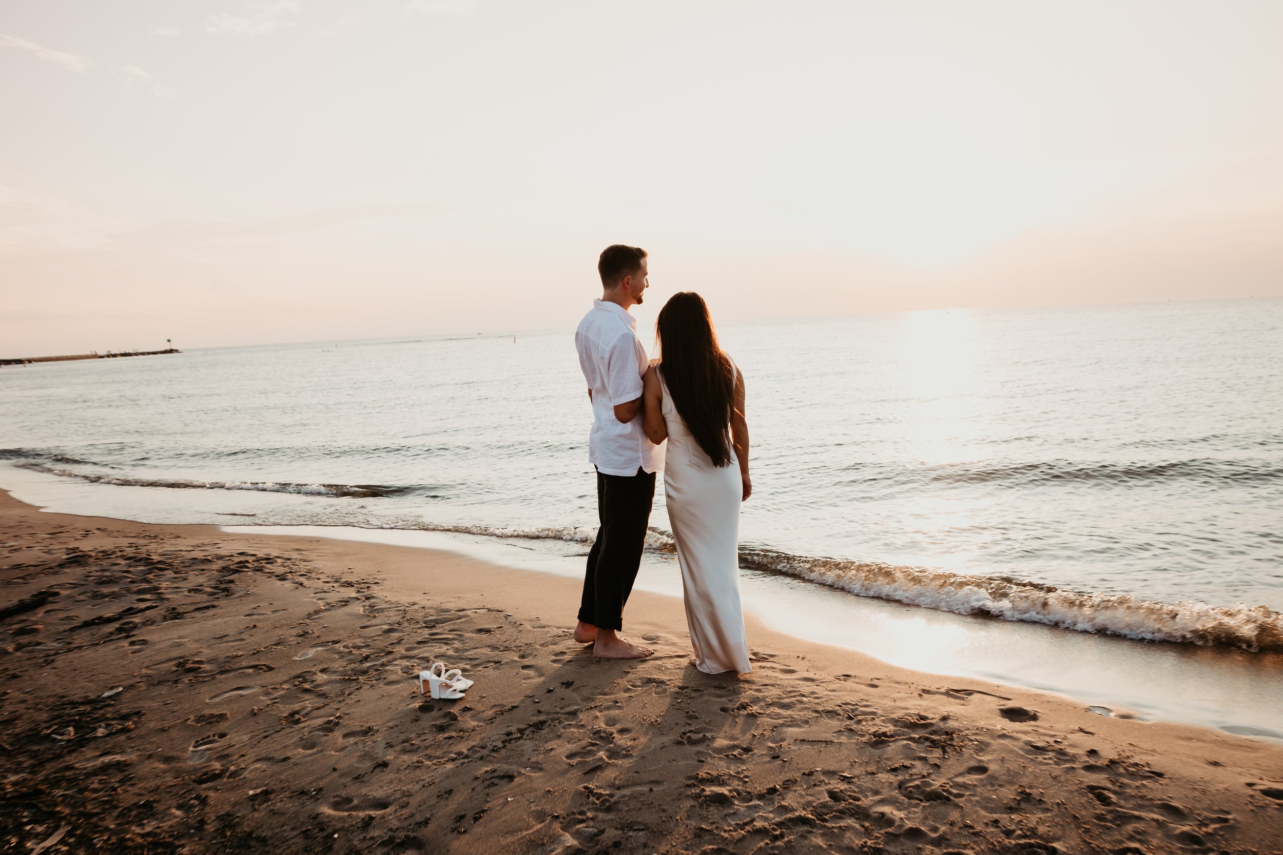 Couple standing together on the shoreline watching the sunset over Lake Michigan during an engagement session at Grand Beach in New Buffalo, Michigan, with the bride-to-be's white heels resting in the sand.