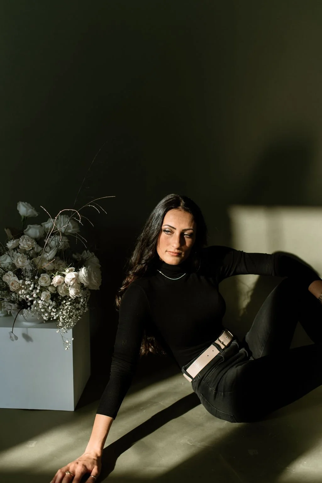 Julia Brown sitting on the floor with her arm resting on the ground, next to a white vase filled with white flowers, illuminated by sunlight casting shadows on a dark wall.