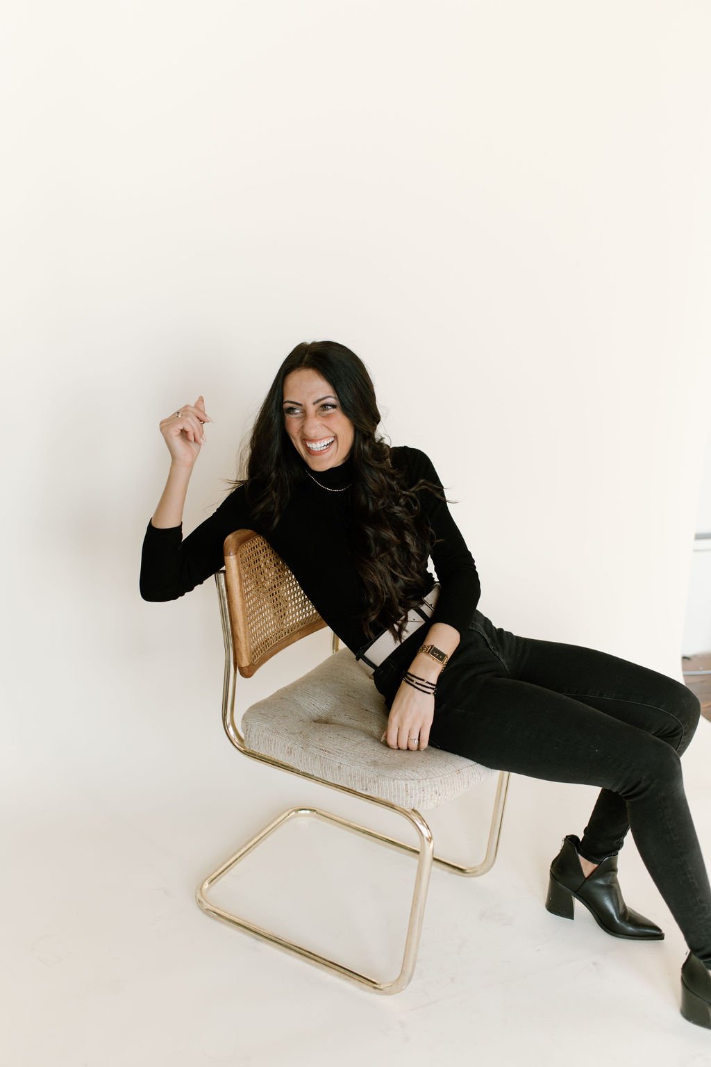 Julia Brown laughing, sitting sideways on a modern chair with a cane back, wearing a black turtleneck and black pants, in a minimalist studio setting.
