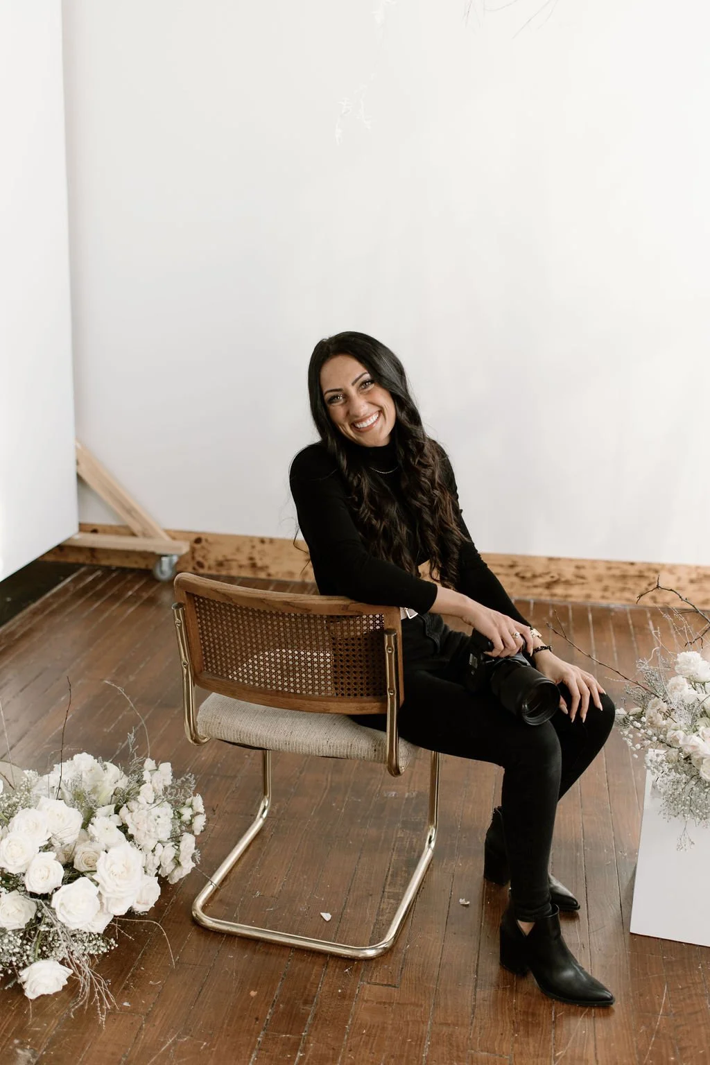 A photo of Julia Brown sitting on the edge of a chair surrounded by floral arrangements on a dark hardwood floor on a white wall background.