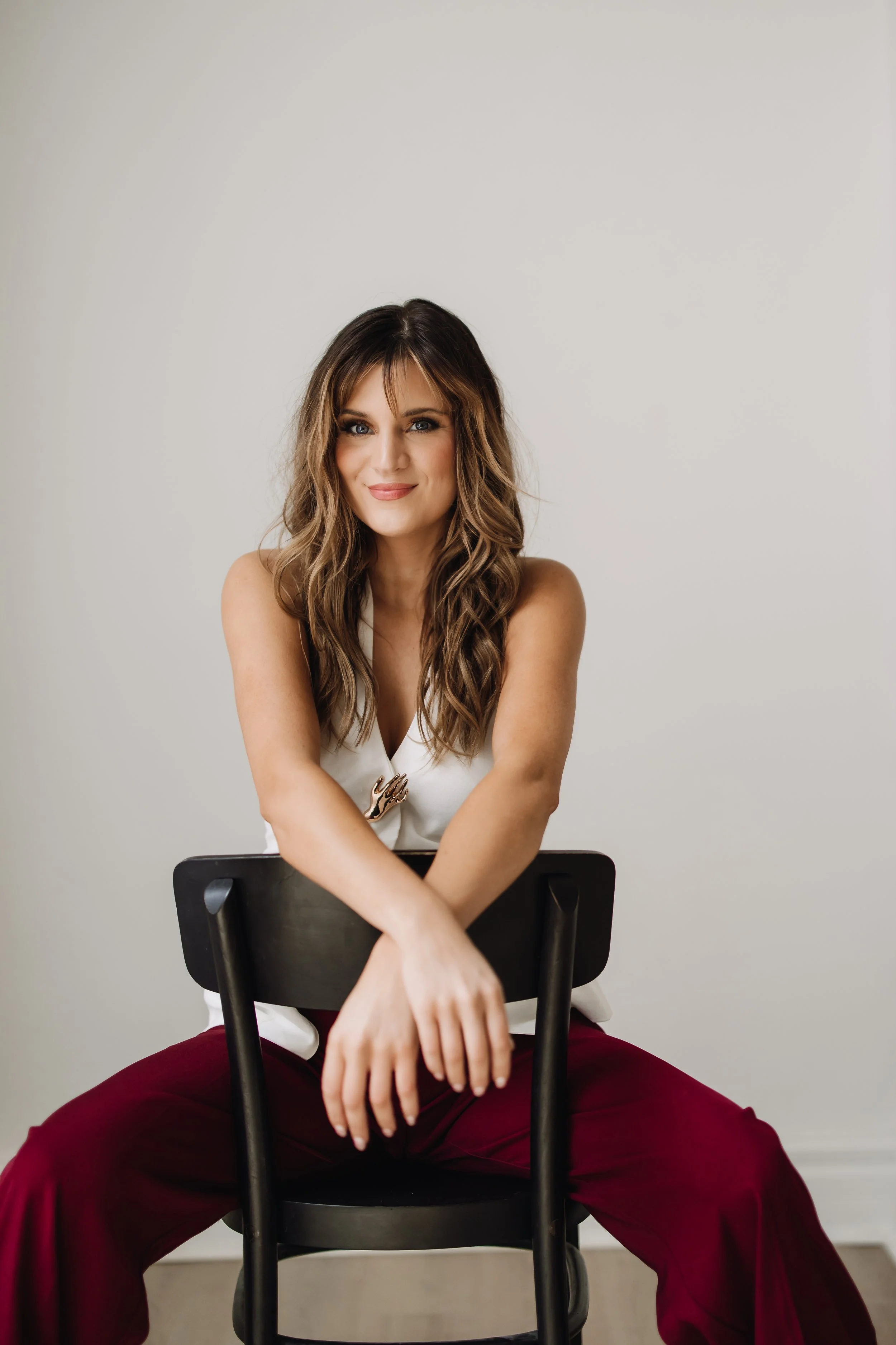 A portrait of A woman with long wavy brown hair and blue eyes sitting on a backwards black chair, wearing a white top and maroon pants, with her arms resting on the back of the chair against a plain white background.