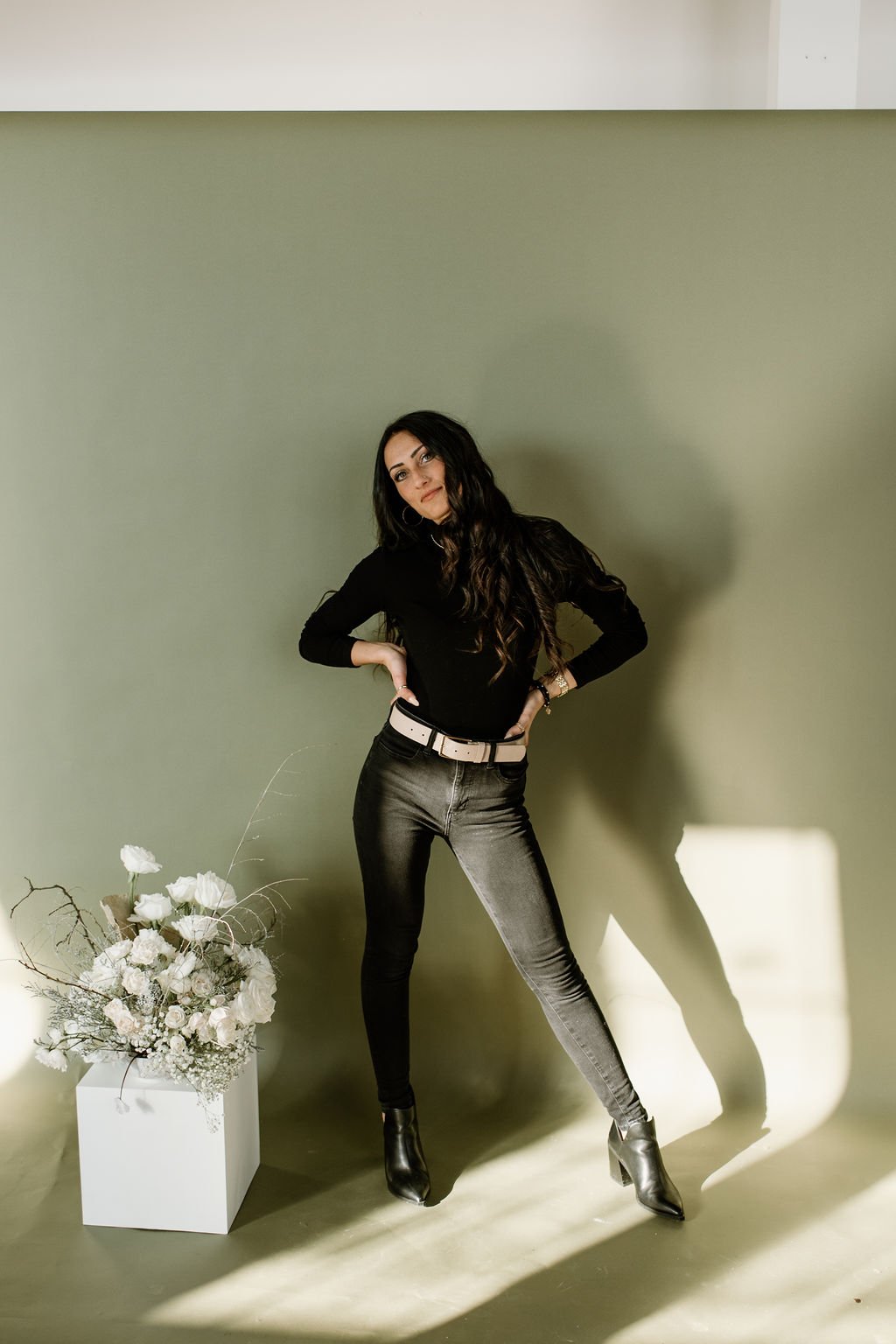 Julia Brown in front of a light green wall posing with her hands on her hips, looking at the camera. There is a white pedestal with a bouquet of white flowers and bare branches to her left, and sunlight casts shadows on the wall behind her.