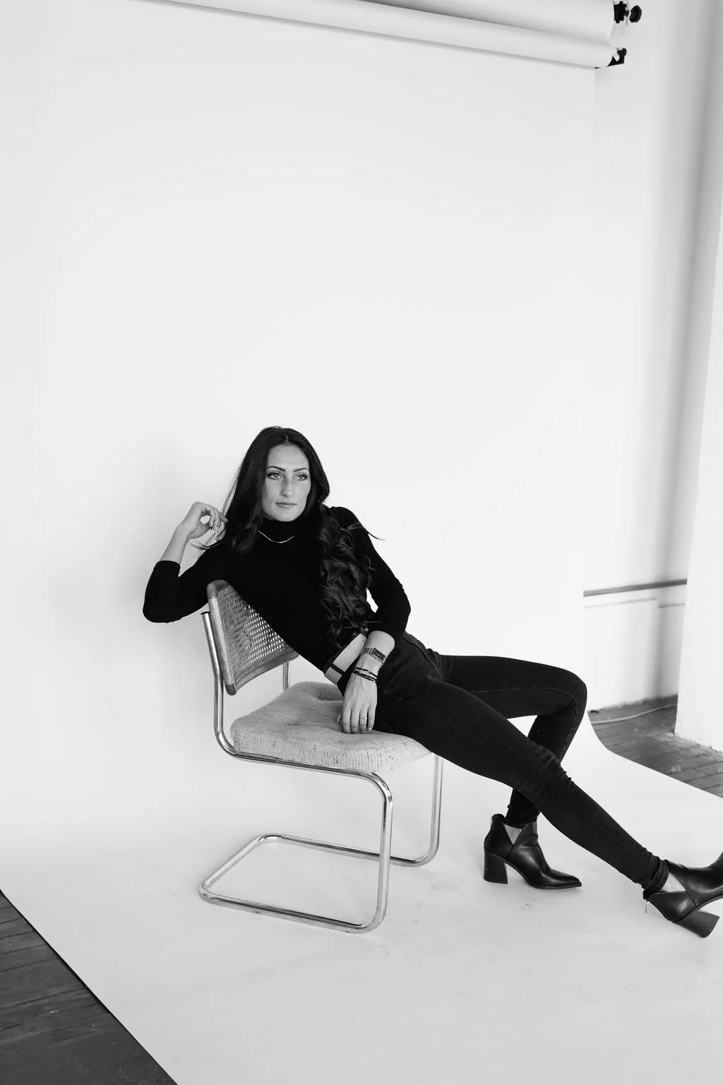 Julia Brown dressed in black, reclining on a chair in a photography studio, with a plain white wall background and studio equipment overhead.