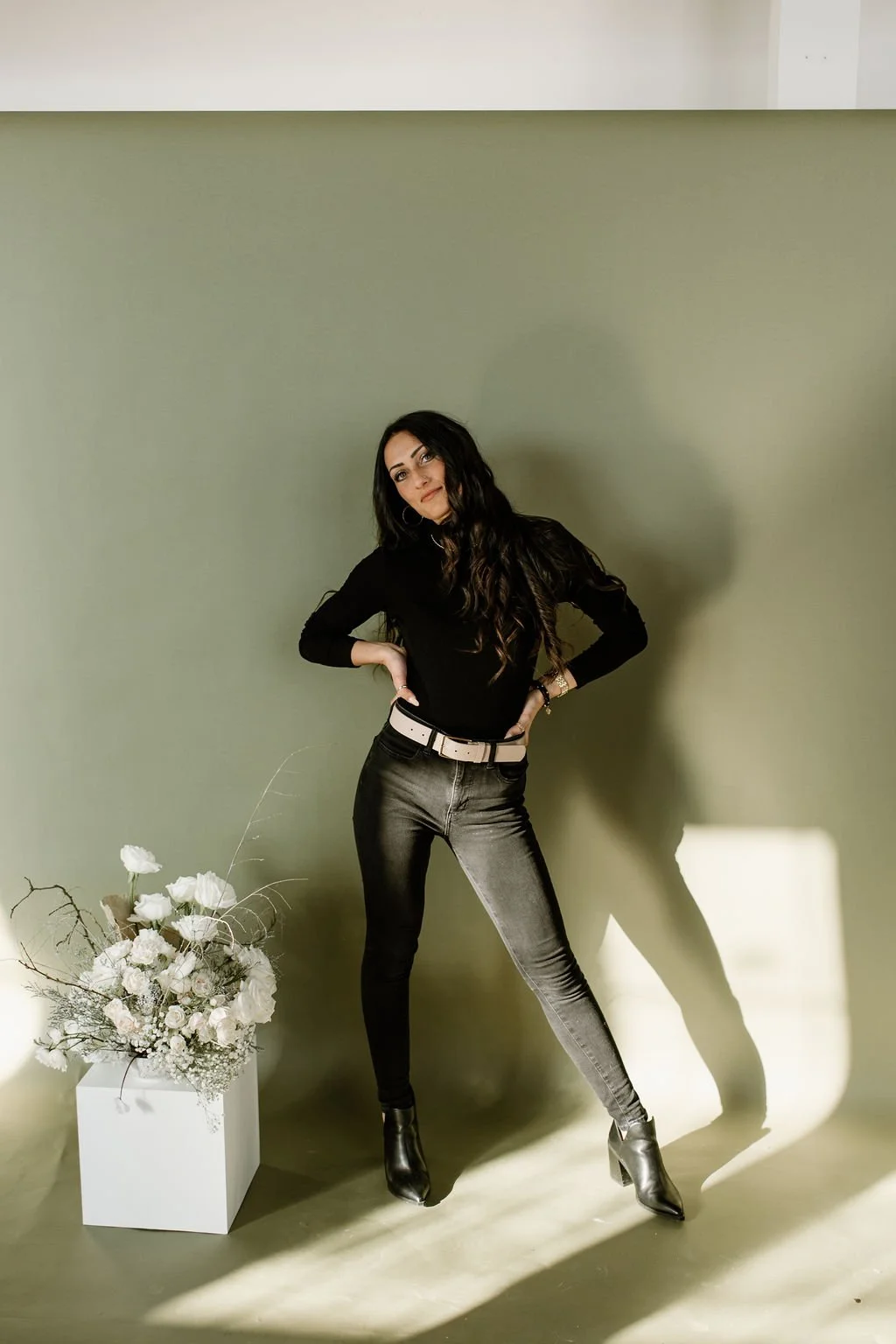 Julia Brown strikes a pose with her hands on her hips in front of an off-white wall next to a white floral arrangement on a decorative cube.