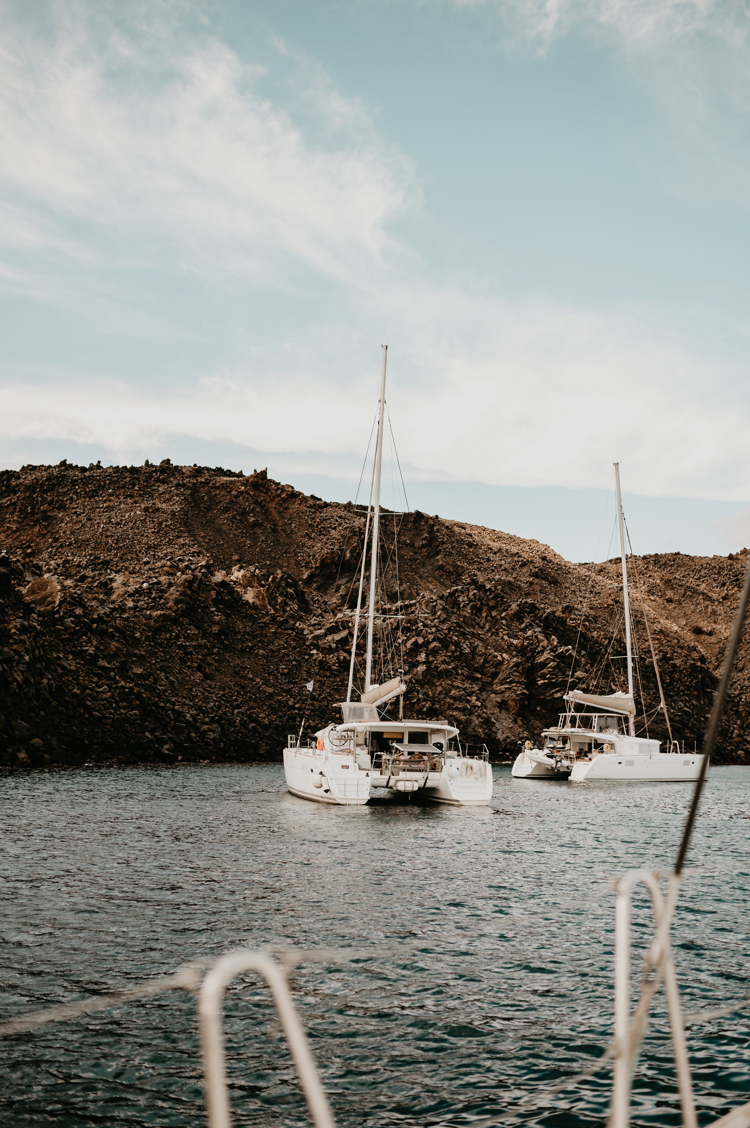 Two sailboats docked in calm water in a Grecian bay with a blue sky in the background.