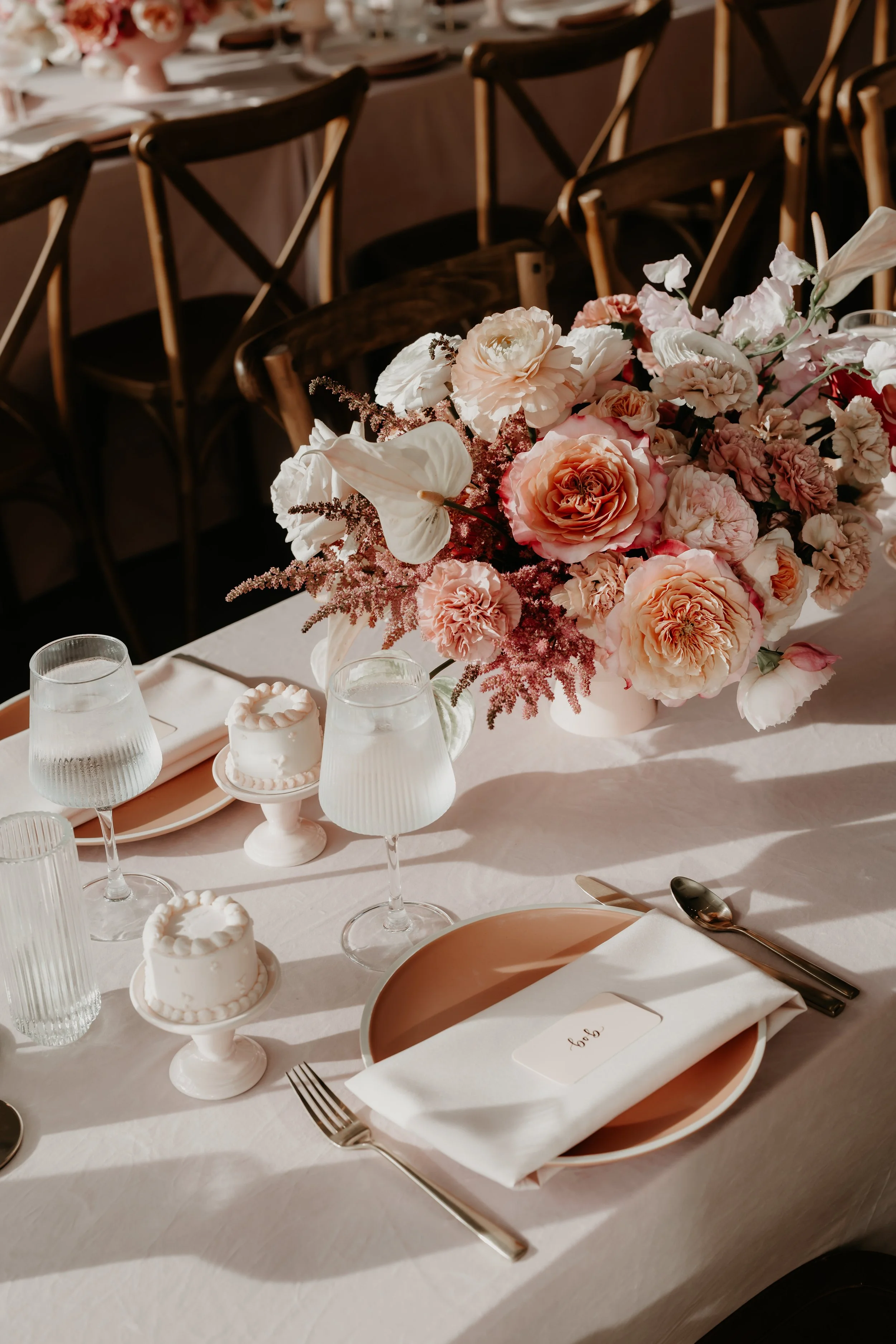 A table at The Wellspring Summer Wedding decorated with a large floral arrangement of pink and white flowers, set with white plates, napkins, silverware, and small cakes on stands.