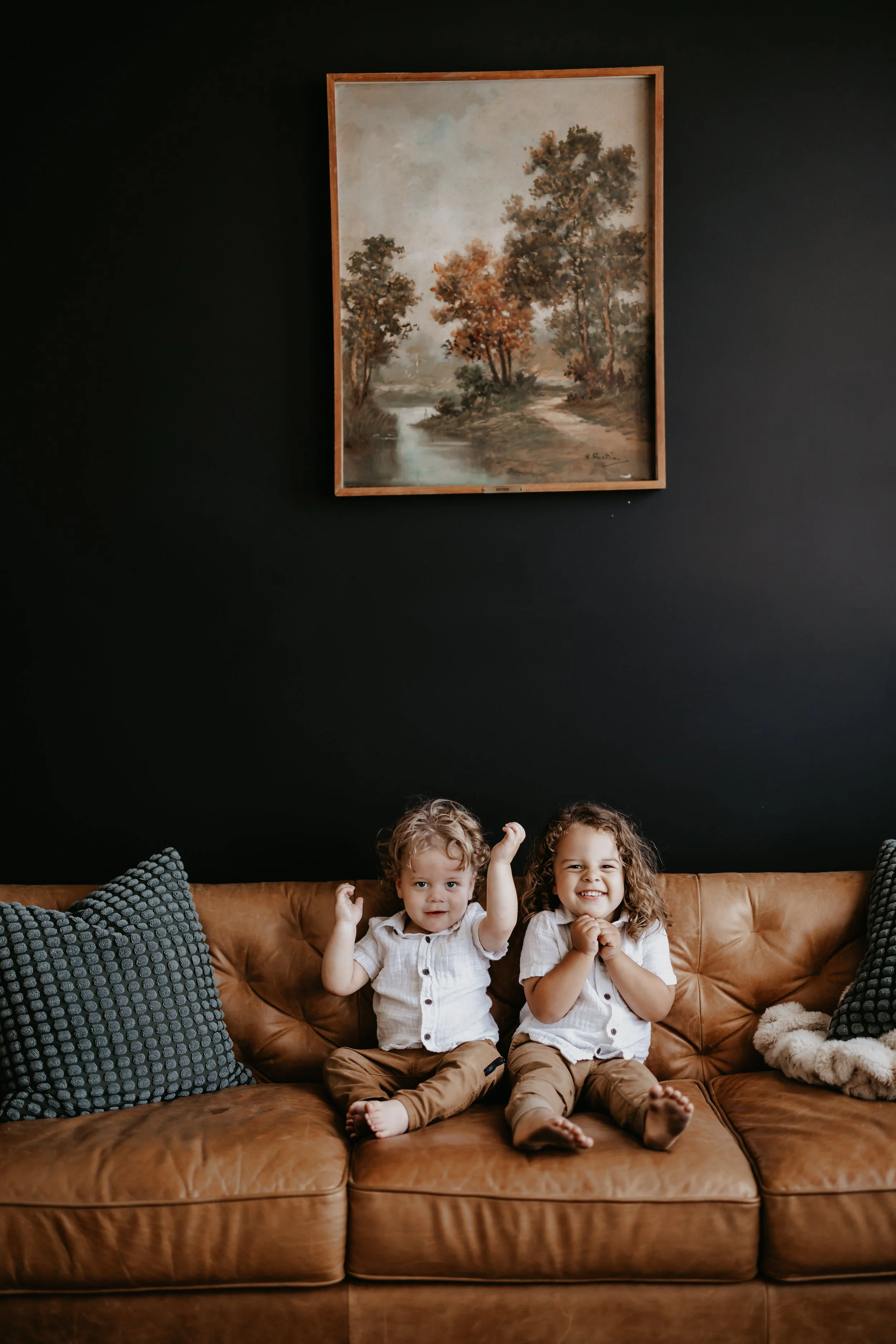 Two young children with curly hair sitting on a brown leather couch, smiling and making gestures, in a living room with a black wall and landscape painting above them.