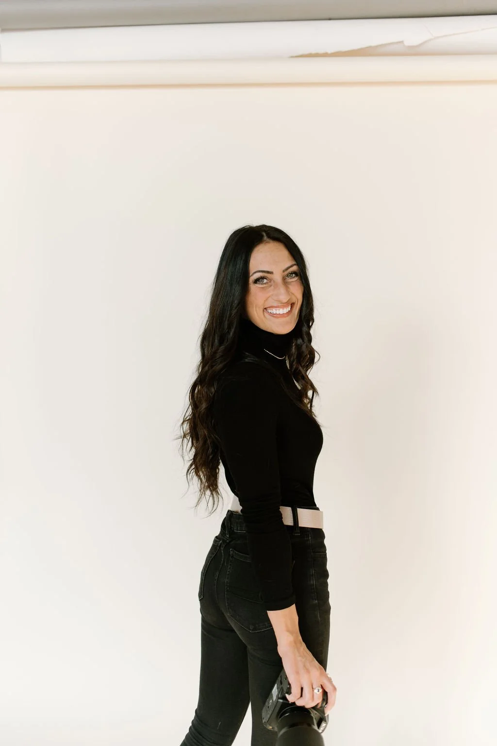 Julia Brown wearing a black top and black jeans, standing against a plain white wall, smiling, holding a camera in her right hand.
