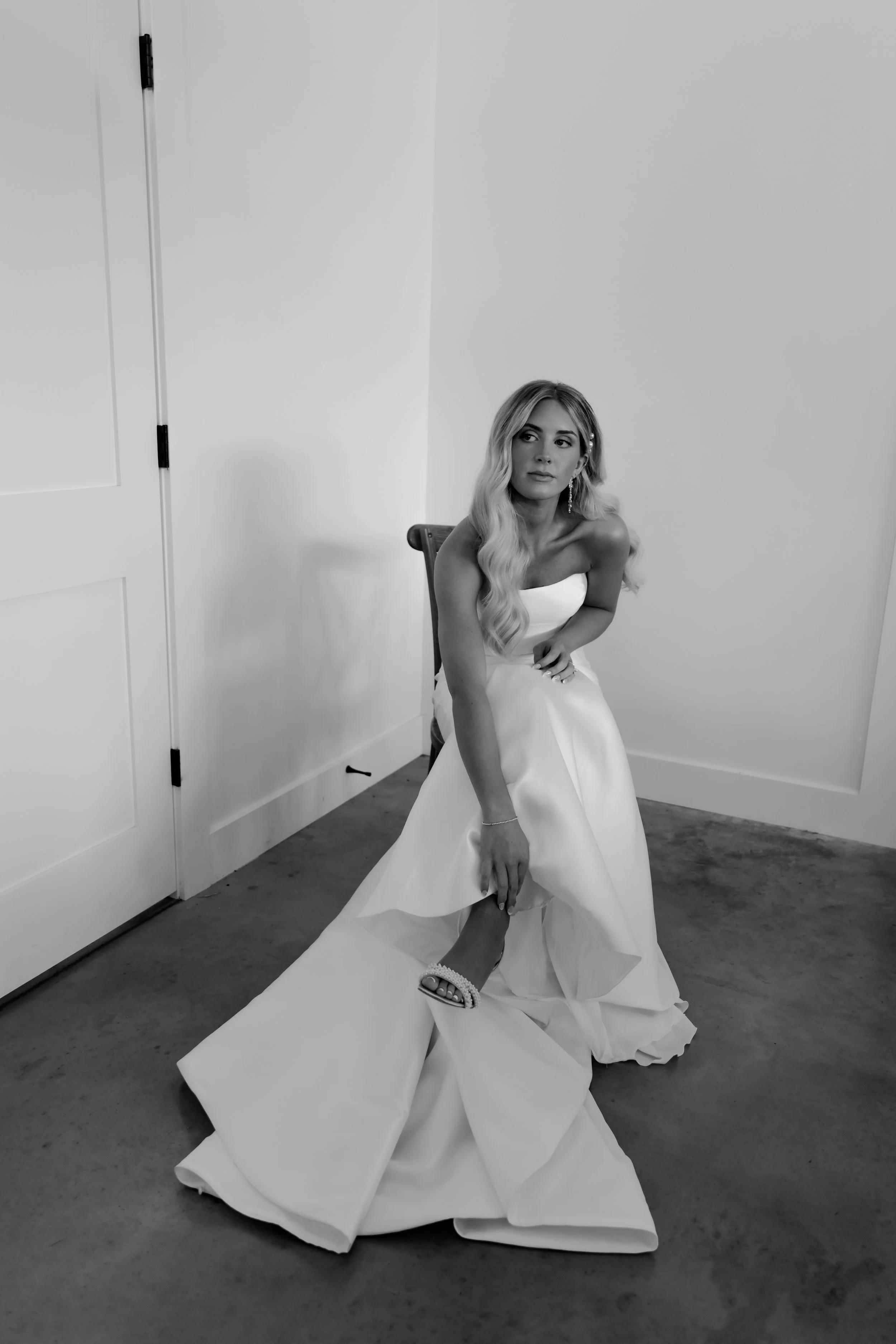 A bride sits in a chair wearing her wedding gown as she reaches down to adjust her high heel shoes. The photo is shot in black and white.
