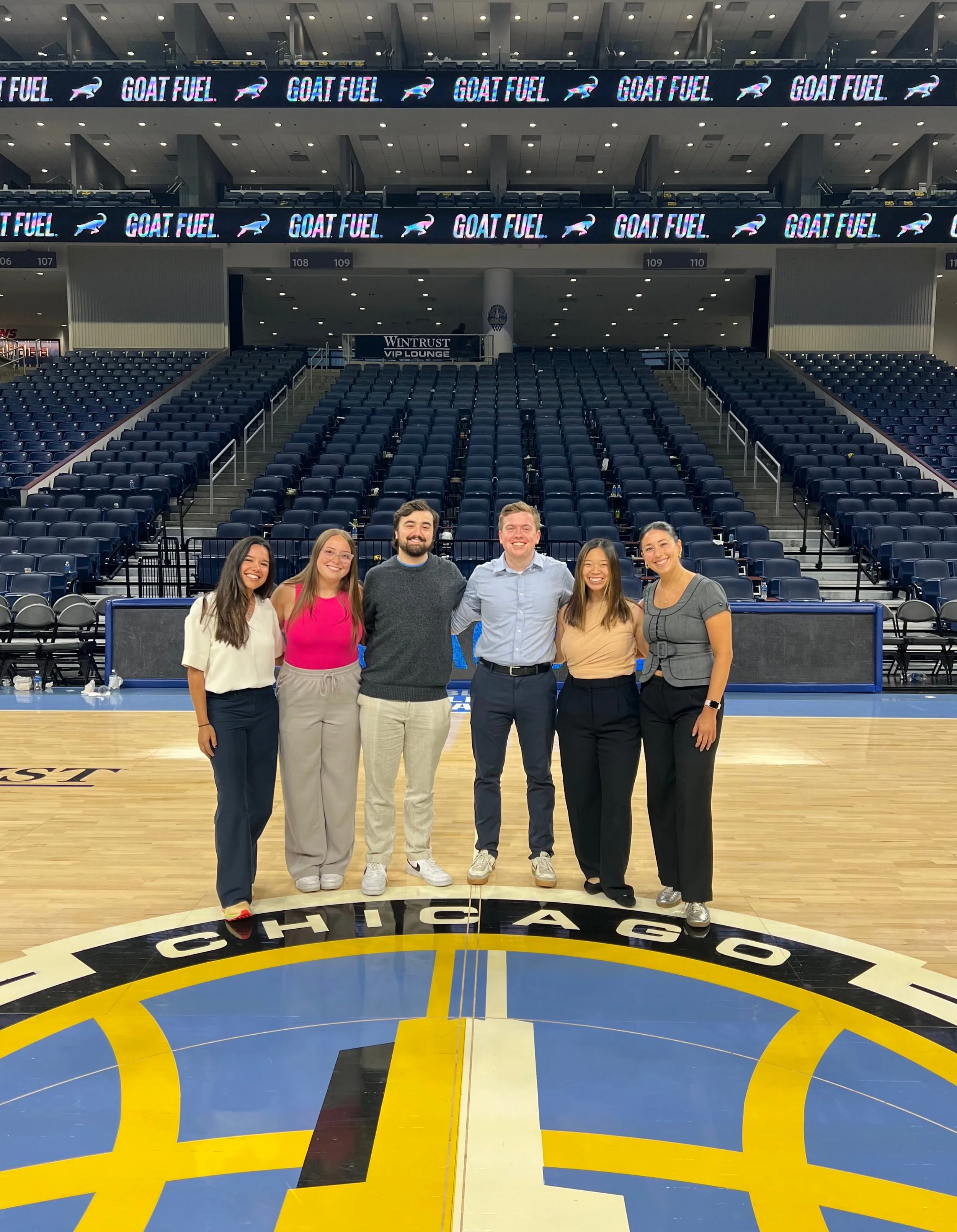 Six people standing on a basketball court in an arena, smiling and posing for a photo, with empty blue seats and digital advertisements in the background.