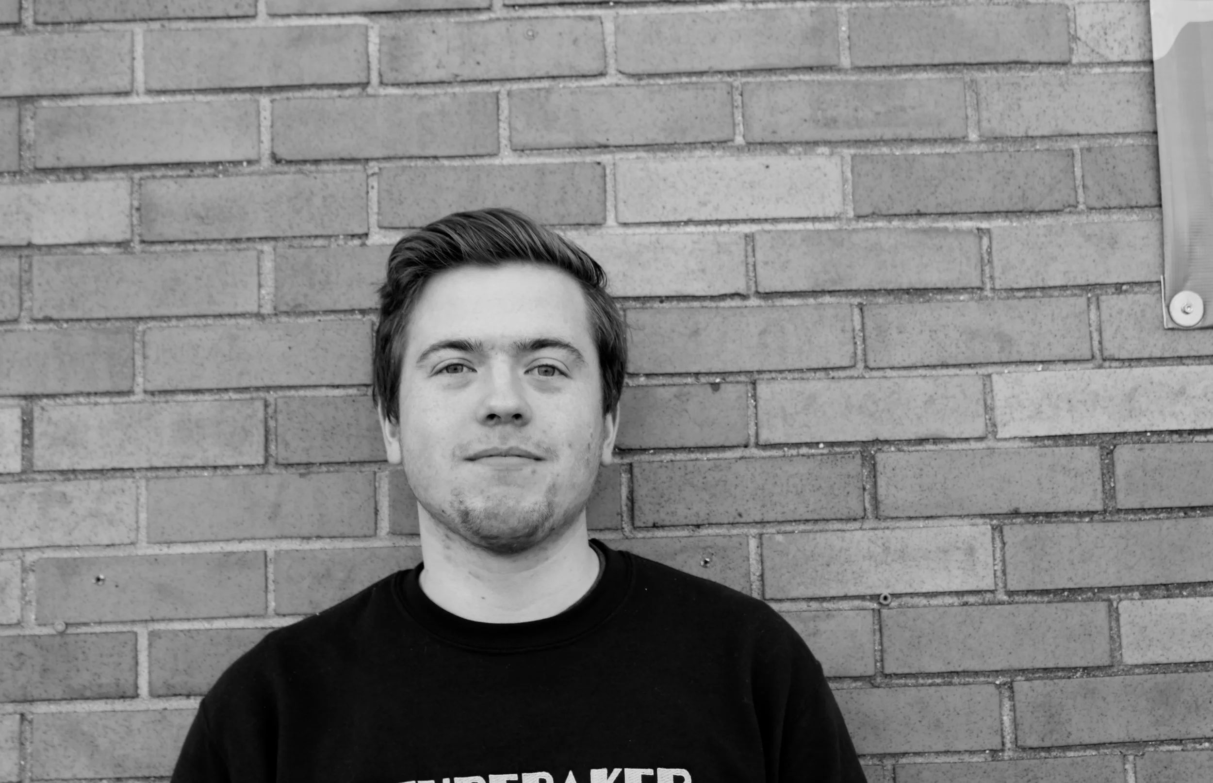Young man with short dark hair and light facial hair standing against a brick wall, wearing a black shirt.