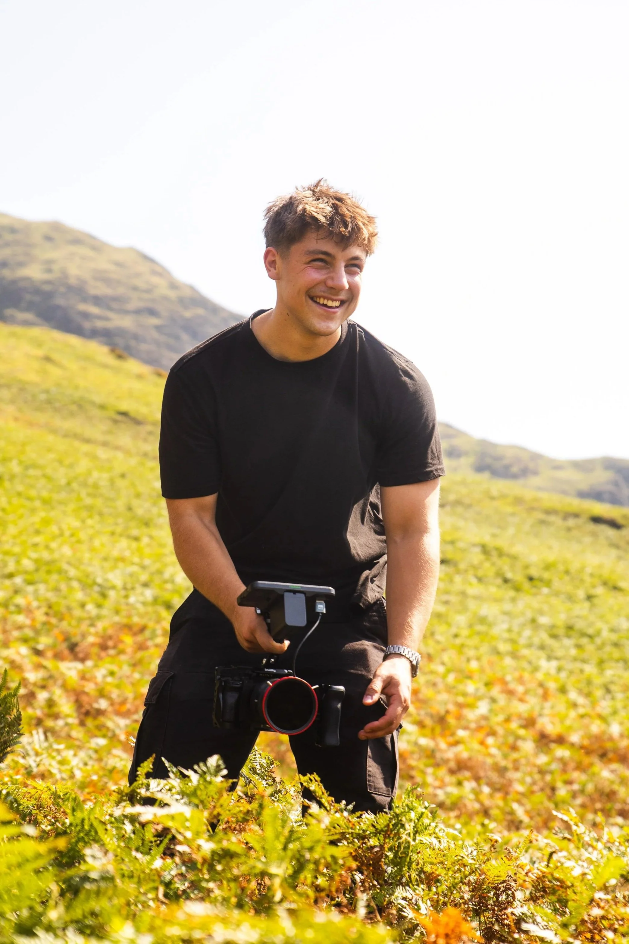A young man in a black t-shirt and shorts holding a camera with a stabilizer, smiling in a green hilly outdoor setting.