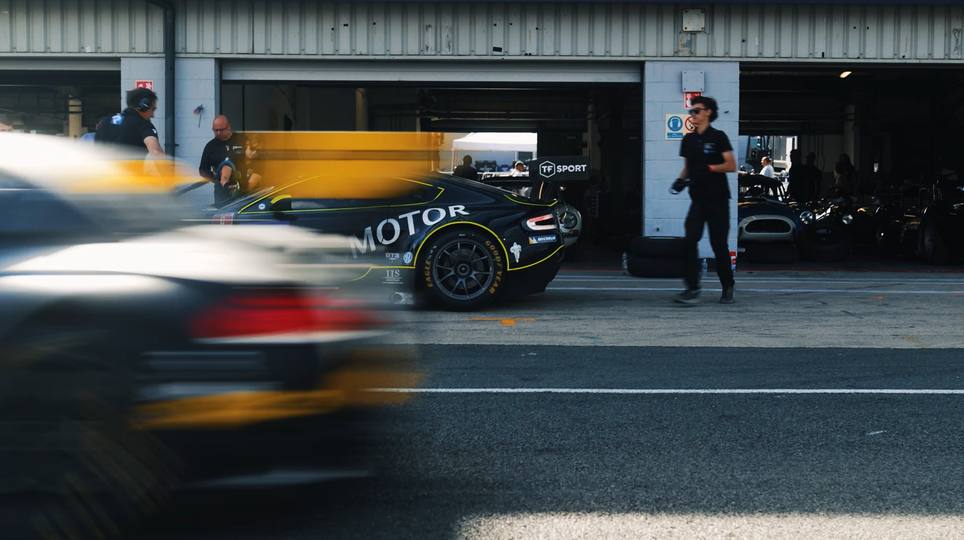 Race car in the pit lane with team members preparing it, while a blurred car passes by in the foreground. The race car is black with yellow accents and has various sponsor logos, including 'MOTOR' and 'GOODYEAR' tires.