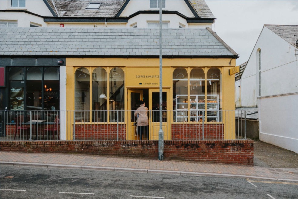 A bakery storefront painted yellow with large windows and a sign reading 'Coffee & Pastry.' A person stands at the door, facing inside. There is a display case with baked goods visible through the window. The bakery is on a street with a brick sidewalk and a metal railing in front.