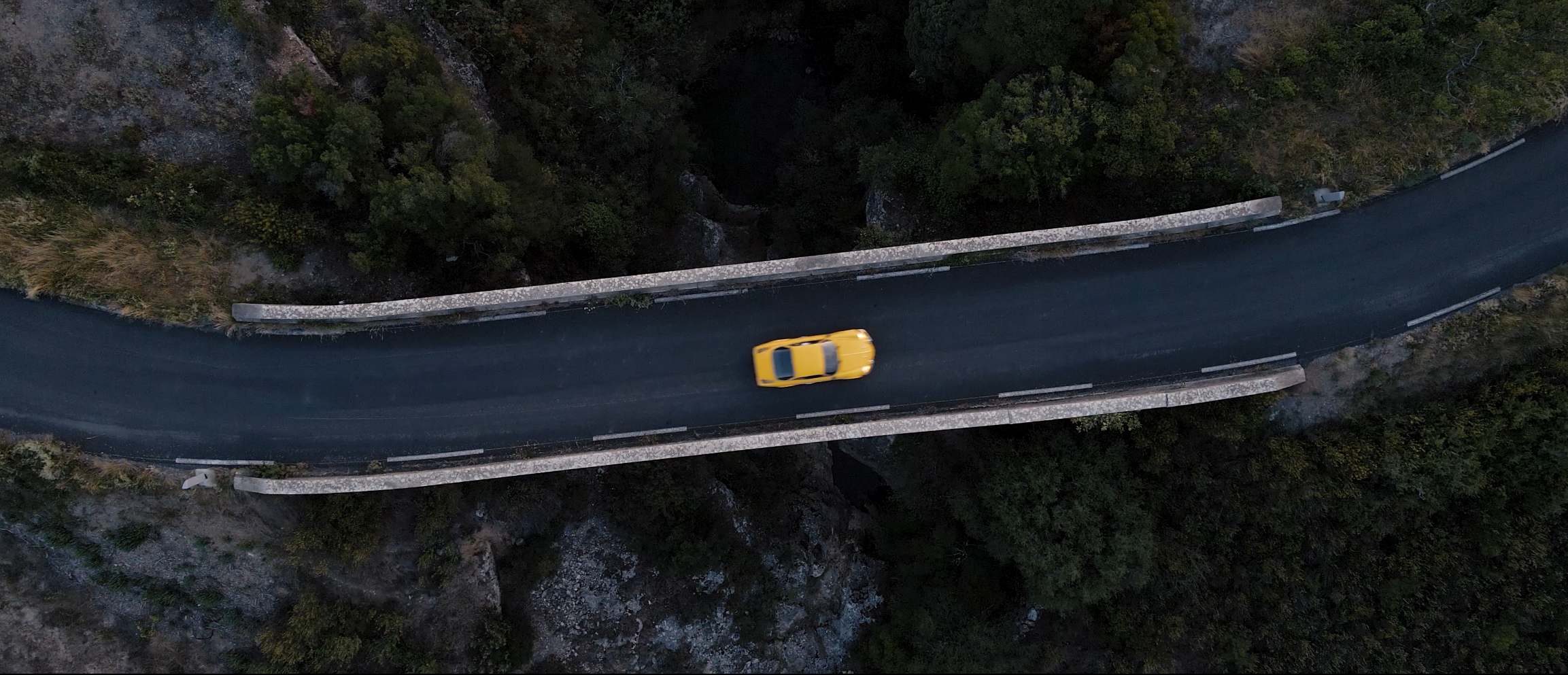 An aerial view of a yellow car driving on a winding mountain road with trees and rocky terrain on either side.