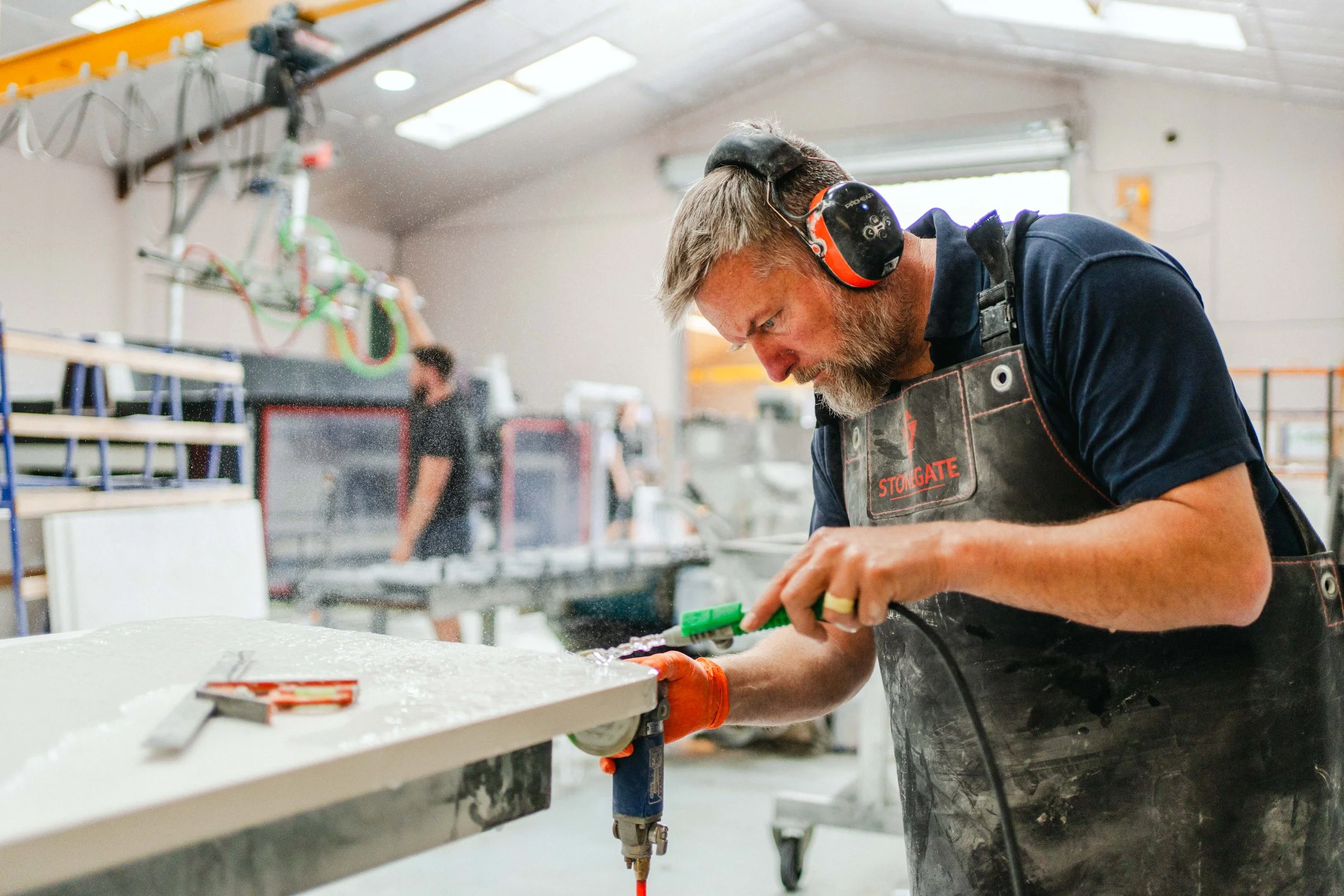 A man working with a power tool in a workshop, wearing safety protective gear including earmuffs and an apron, with other workers in the background.