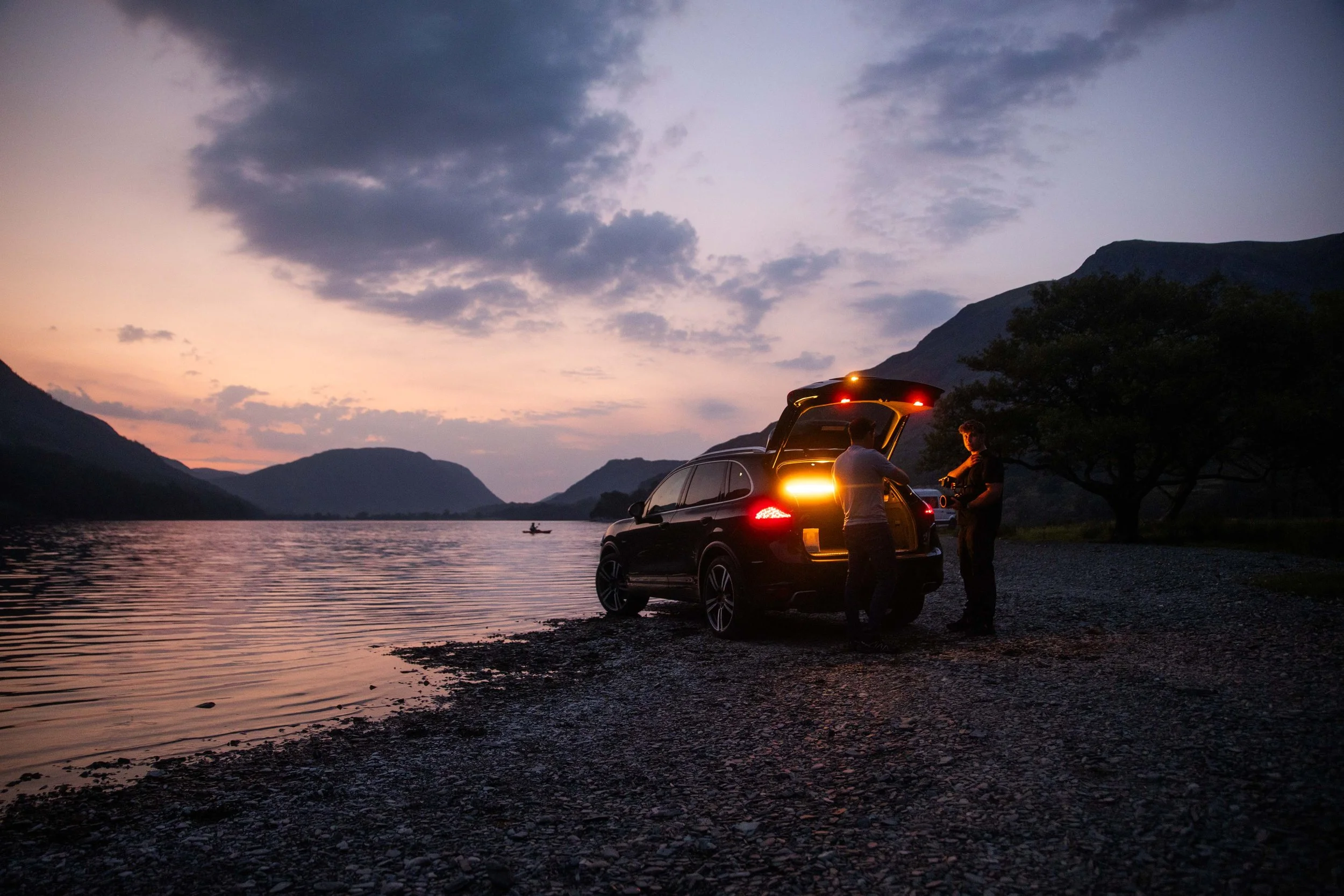 Two people standing next to a black SUV with its trunk open by a lake at sunset, with mountains and sky in the background, and a person in a small boat on the water.