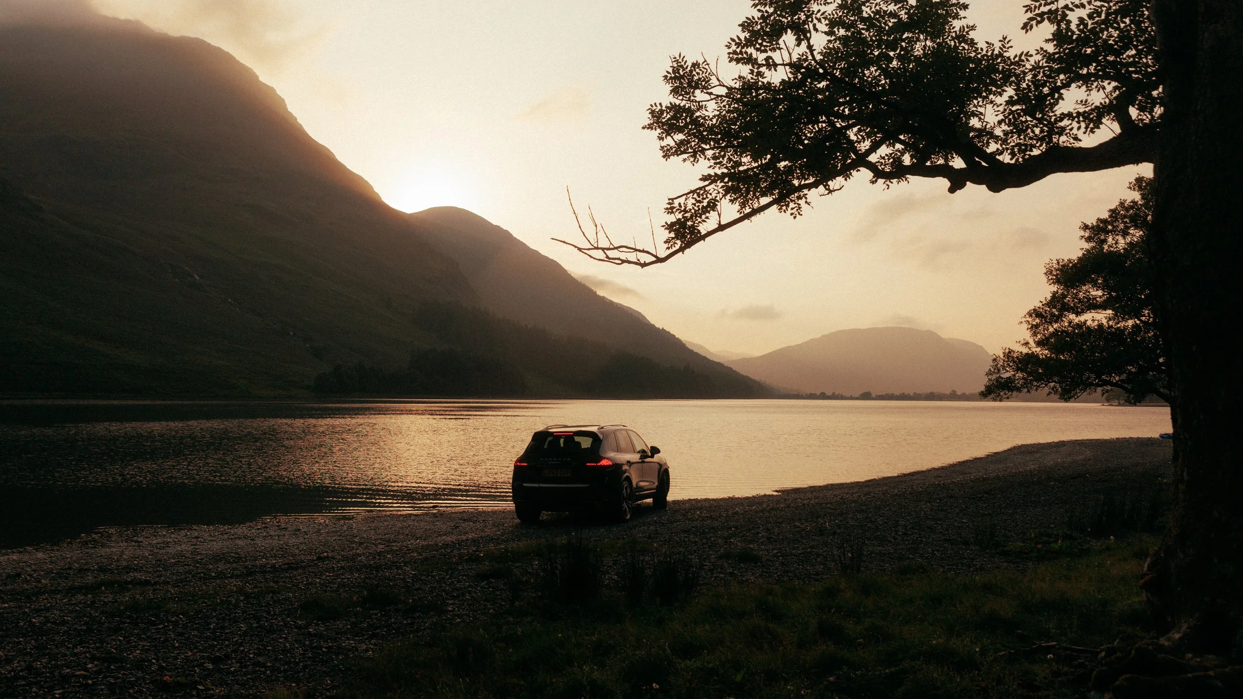 A car parked on a pebbled shoreline next to a calm lake or river, with mountains and a setting sun in the background, a large tree with overhanging branches on the right side of the image.