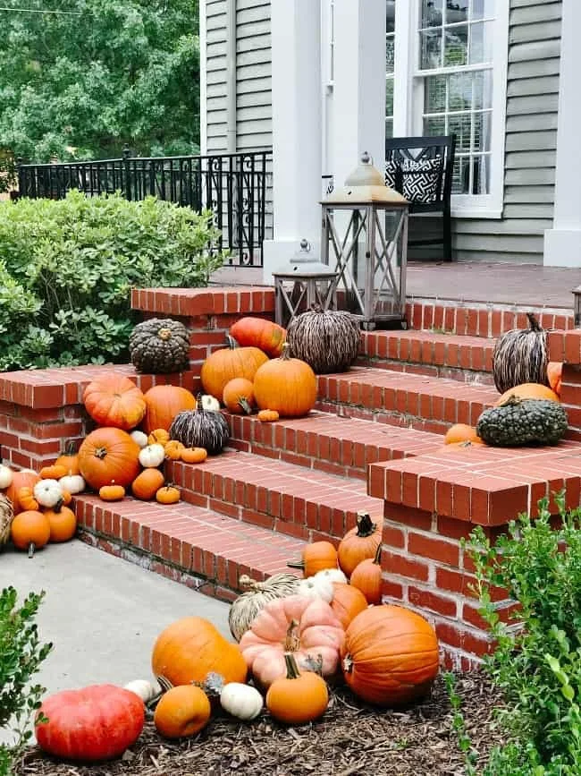 Fall display of pumpkins and gourds on brick steps leading to a porch with a black chair and lanterns.