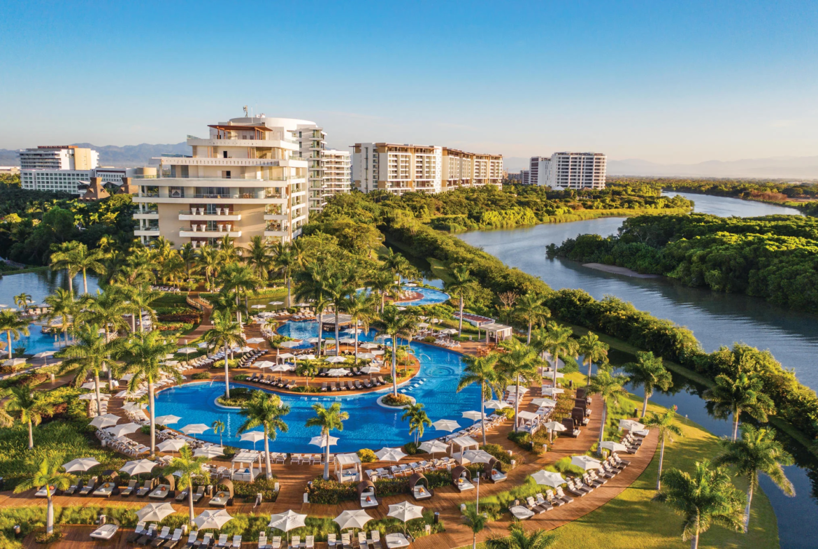 Luxury resort with multiple swimming pools, palm trees, sun loungers with umbrellas, and modern high-rise buildings next to a river amid greenery, under a clear blue sky.