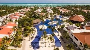 Aerial view of a resort with multiple swimming pools, surrounded by palm trees, pathways, and buildings with red roofs, near a beach with ocean in the background.