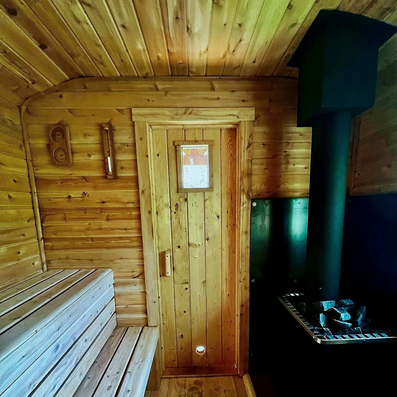 Interior of a custom wood-fired sauna by Hot Bird Saunas with tongue and groove walls, bench seating, a small window, and a black stone heater.