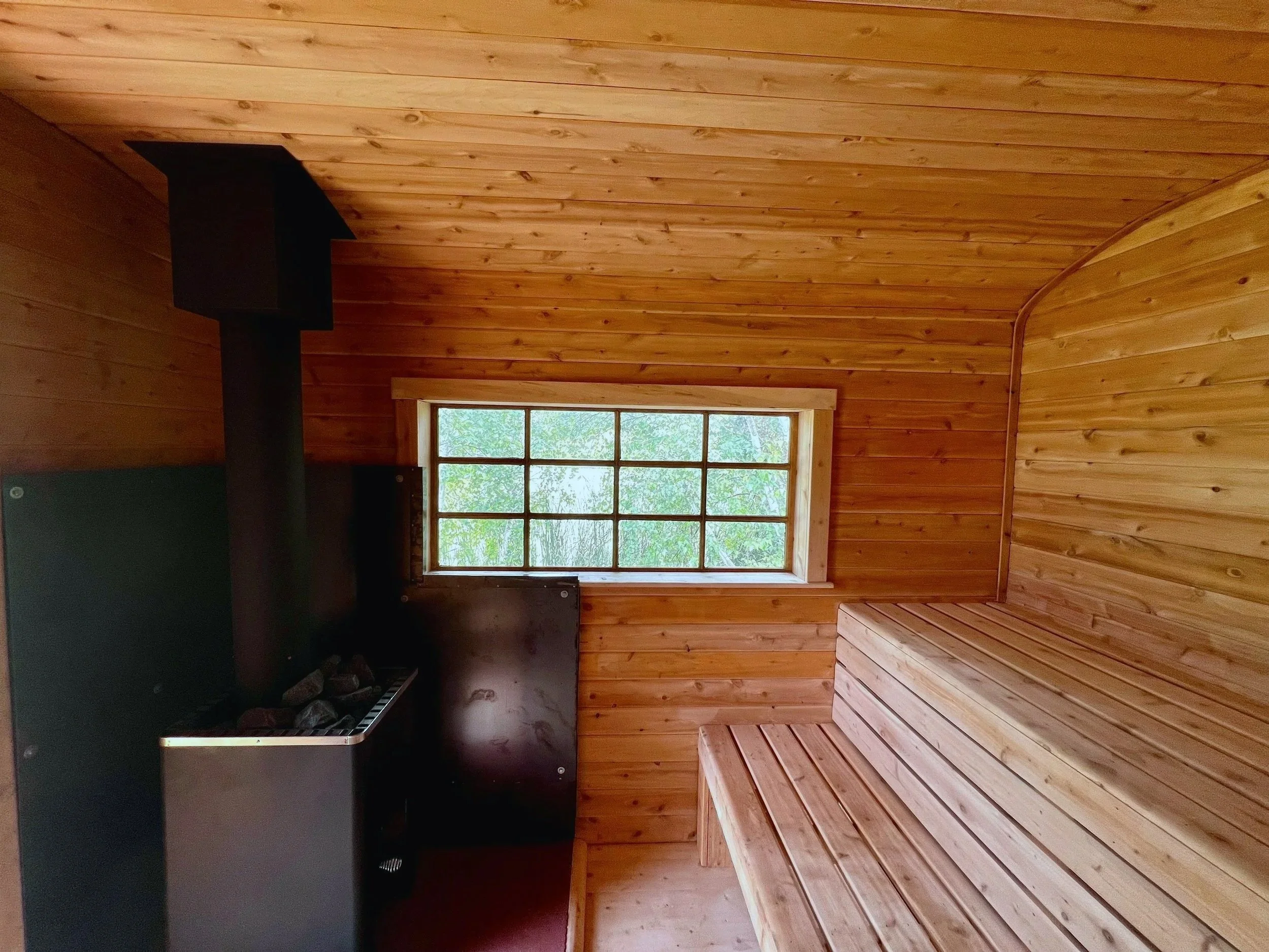 Interior of a custom wood-fired sauna by Hot Bird Saunas with raised bench seating, a window overlooking trees, and a black wood-fired heater.
