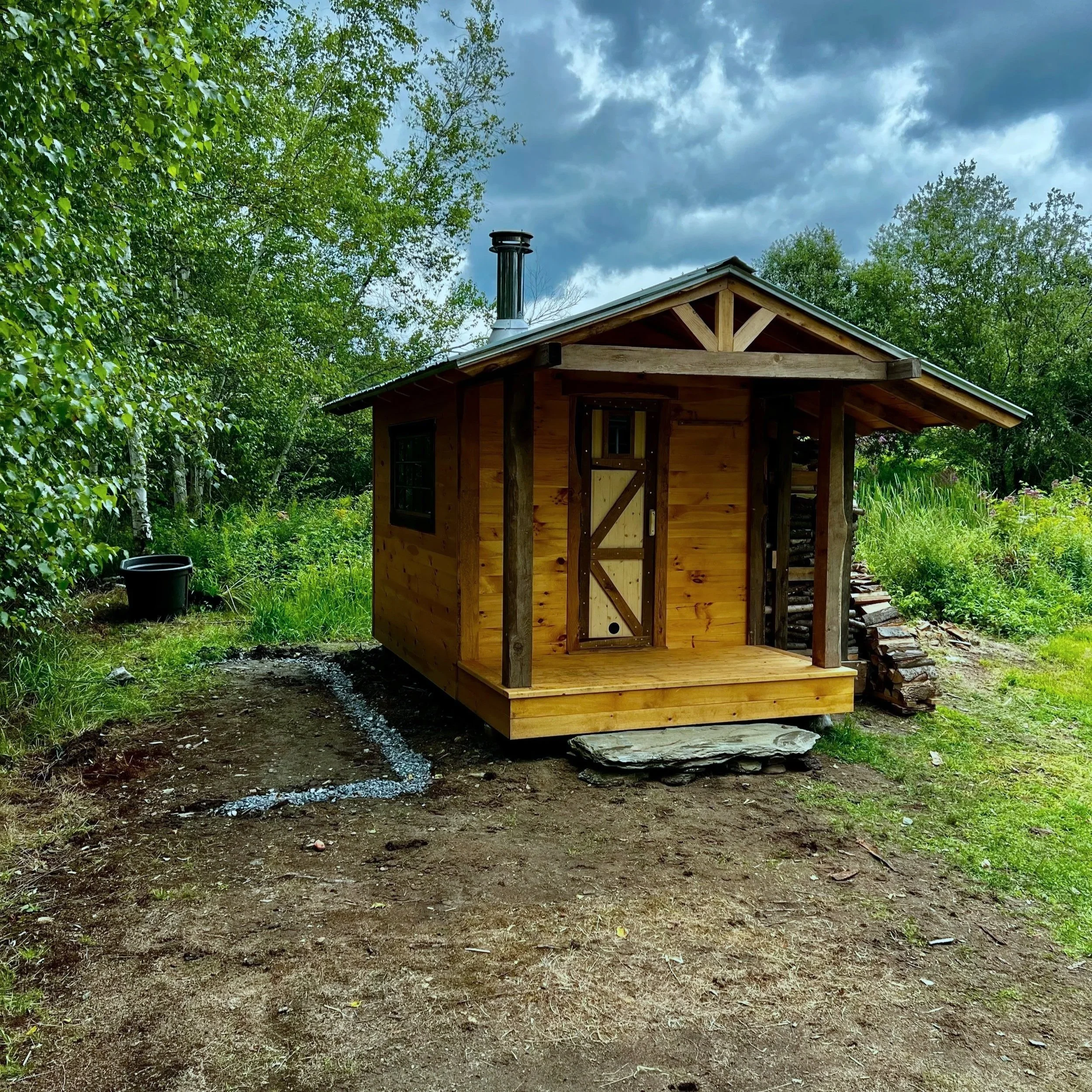 Custom outdoor sauna with covered entry porch by Hot Bird Saunas, set along a dirt path and surrounded by trees and greenery.