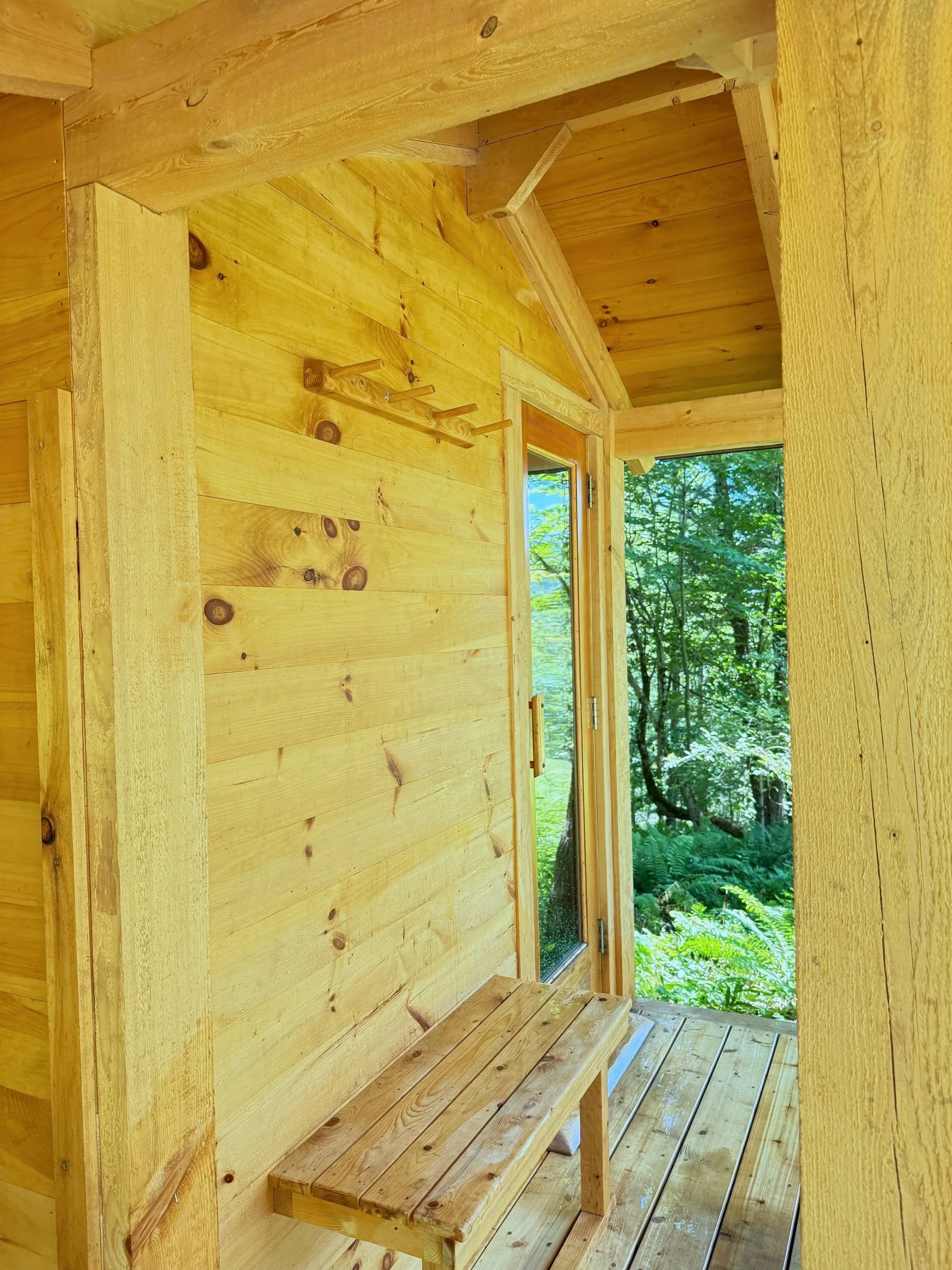 Interior entry area of a custom sauna with cedar tongue and groove, a bench and wall hook, and an open door looking out onto surrounding greenery.