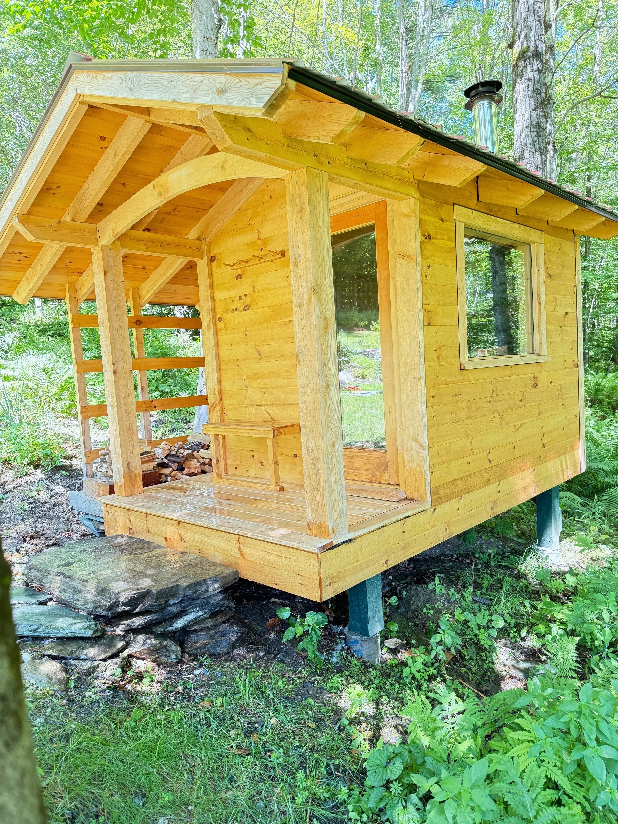 Custom outdoor sauna by Hot Bird Saunas with a covered porch, elevated on stilts and surrounded by trees and green foliage.