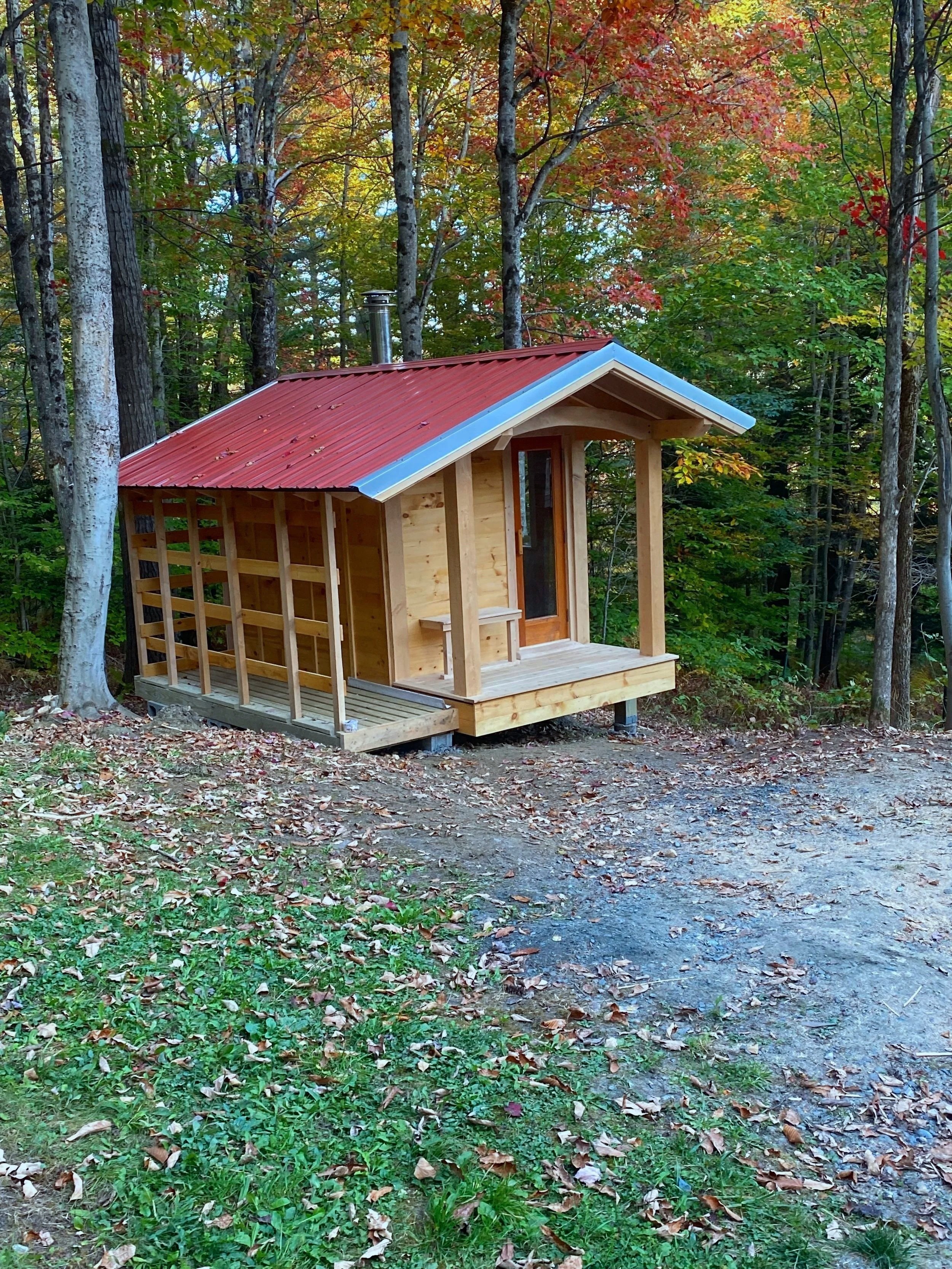 Custom outdoor sauna with a red metal roof, set among autumn trees with colorful foliage.