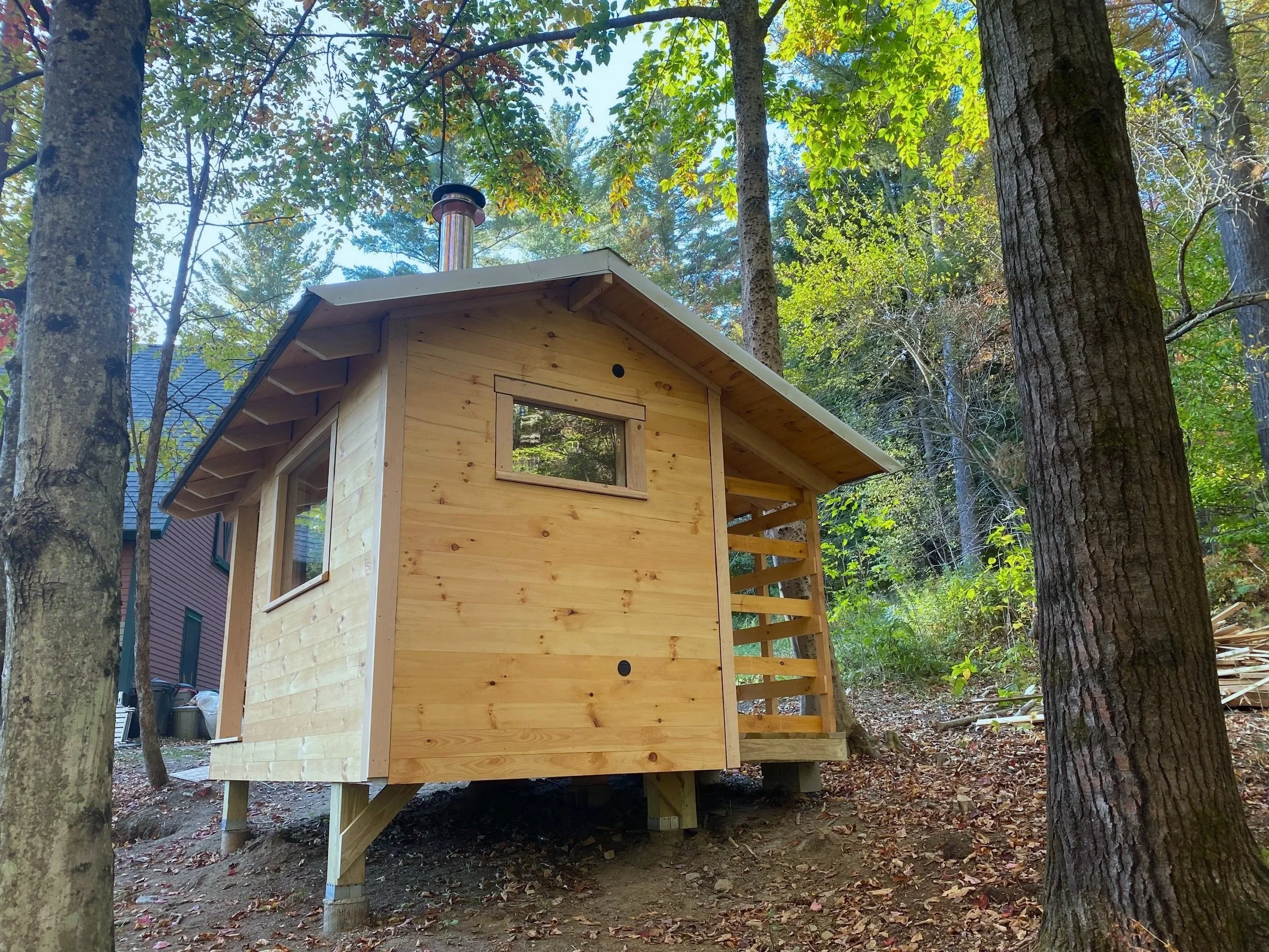 Custom outdoor sauna with a gabled metal roof, small window, and partial deck, set among trees and fallen leaves.