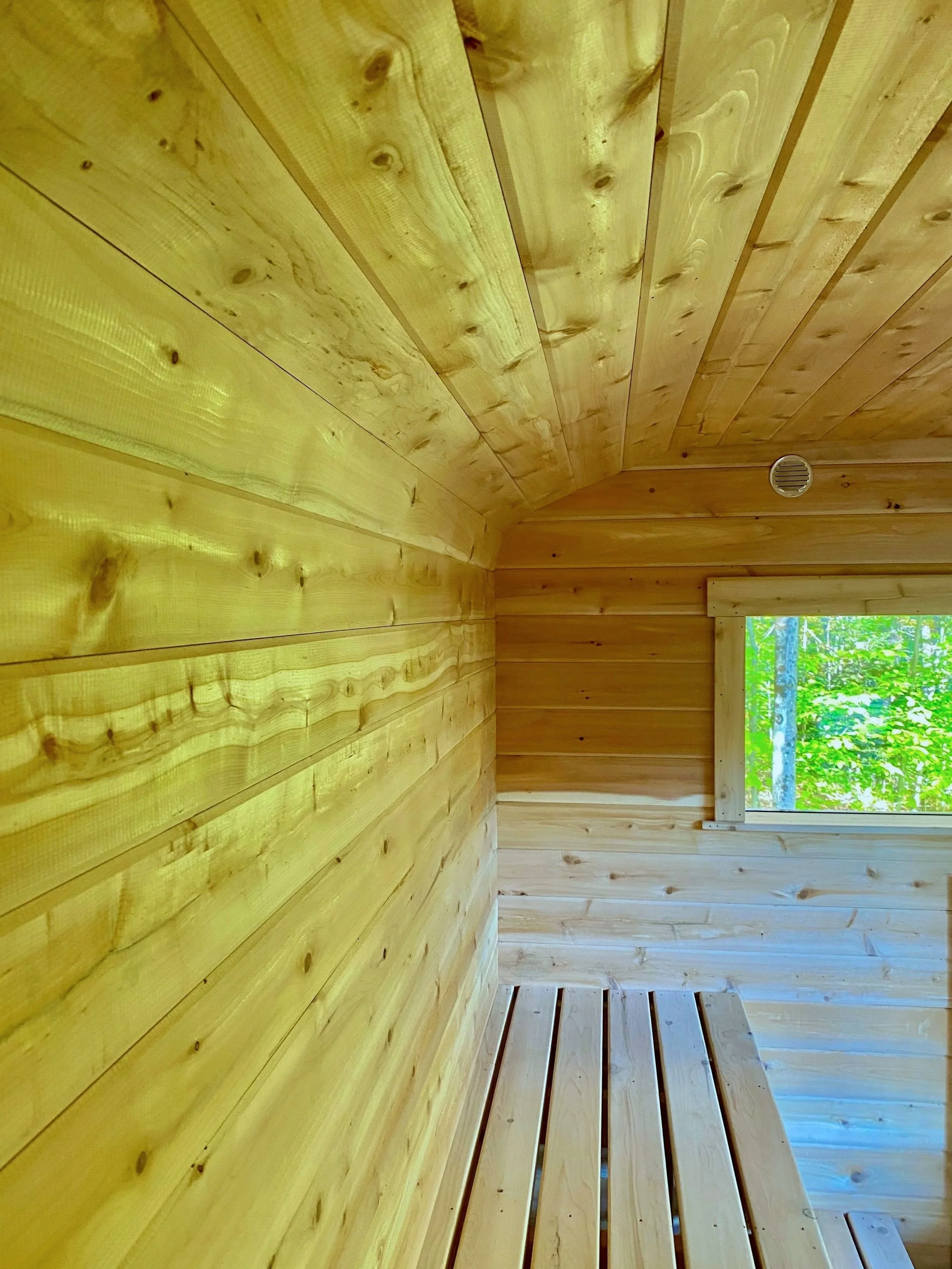 Interior of a custom sauna with smooth, light-colored cedar walls and ceiling, a built-in wooden bench, and a window looking out onto greenery.