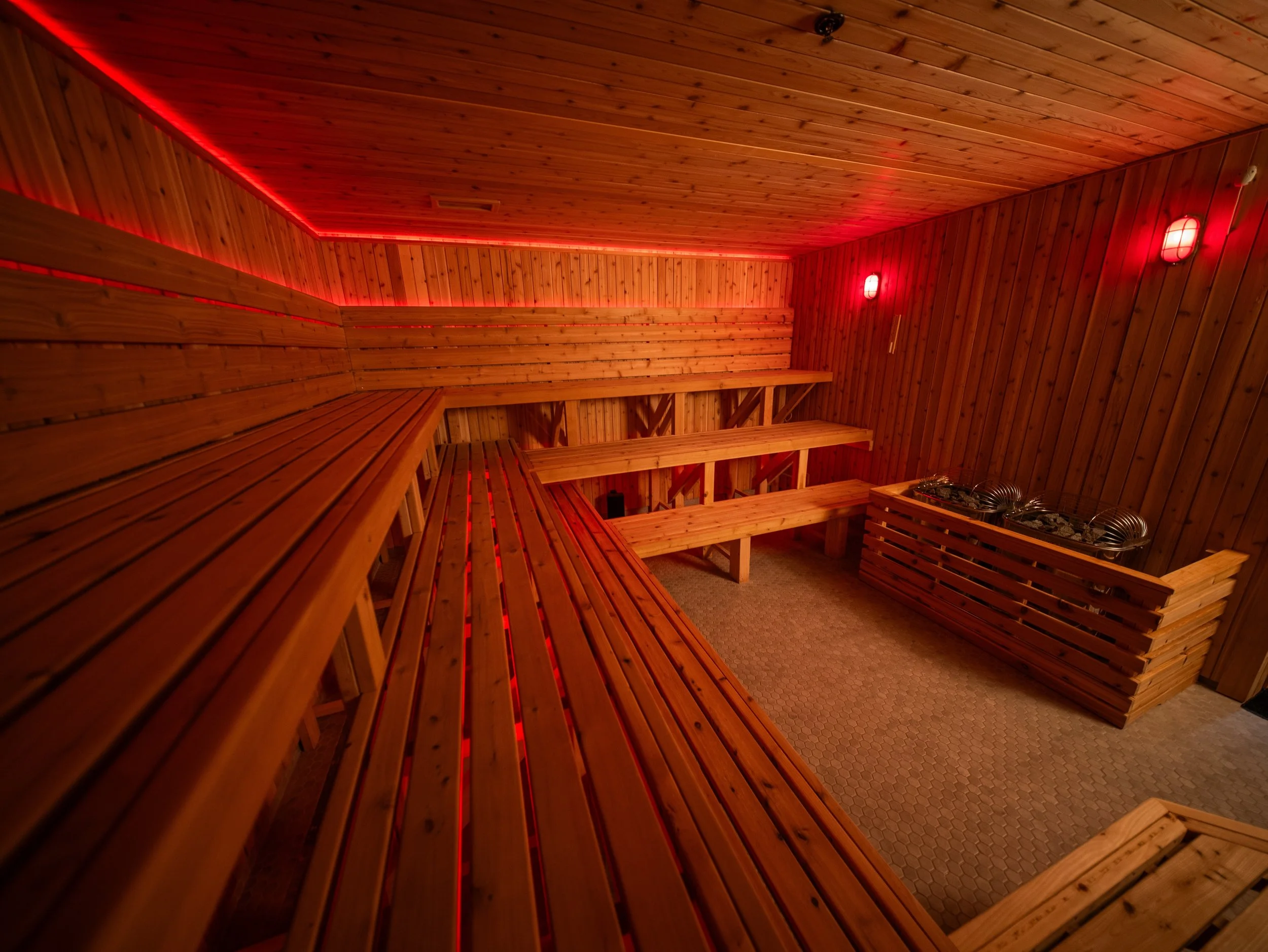 Custom sauna interior by Hot Bird Saunas with tiered cedar seating, a traditional stone heater, and ambient red LED lighting.