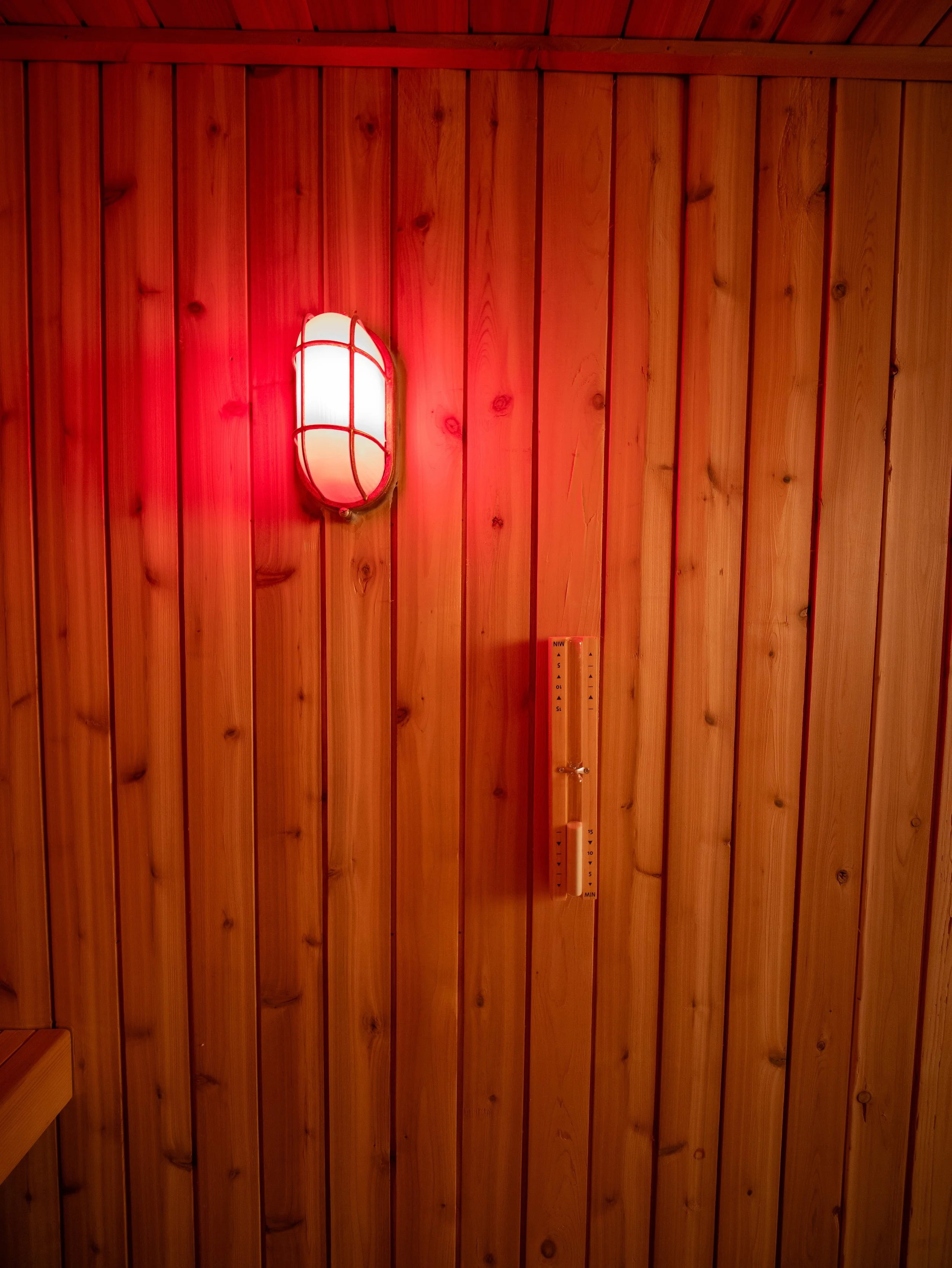 Wall detail inside a custom sauna showing wood paneling, a red light fixture, and a traditional wooden thermometer.