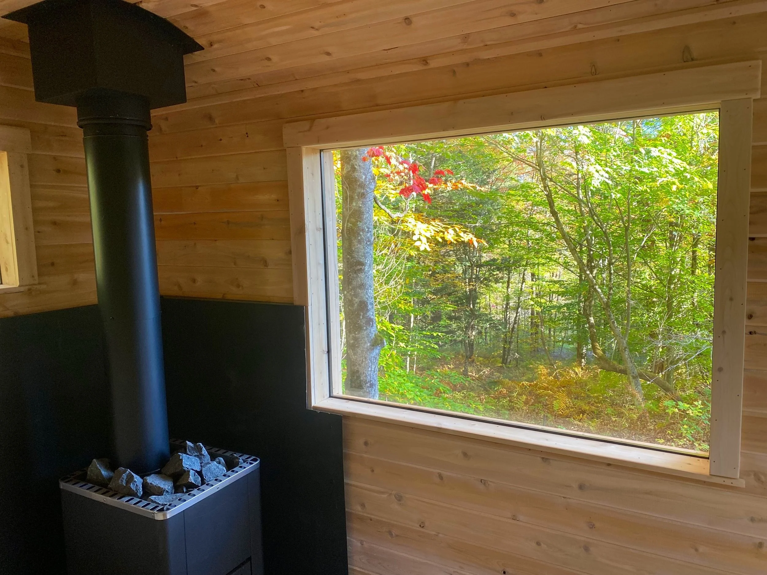 Interior of a custom wood-fired sauna with cedar tongue and groove walls, a large picture window overlooking trees, and a stone-topped wood stove in the corner.