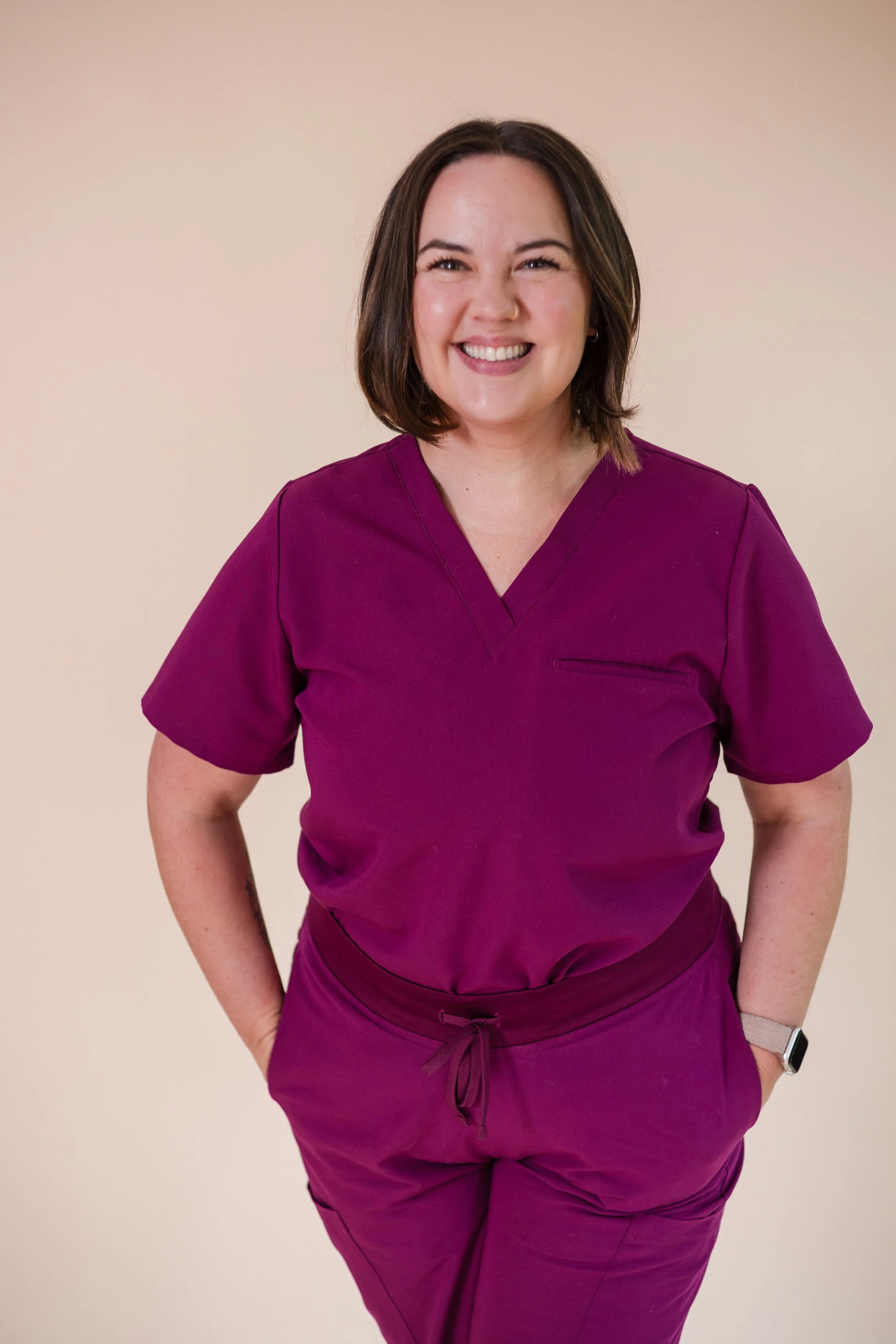 A woman in purple medical scrubs smiling with her hands in her pockets.