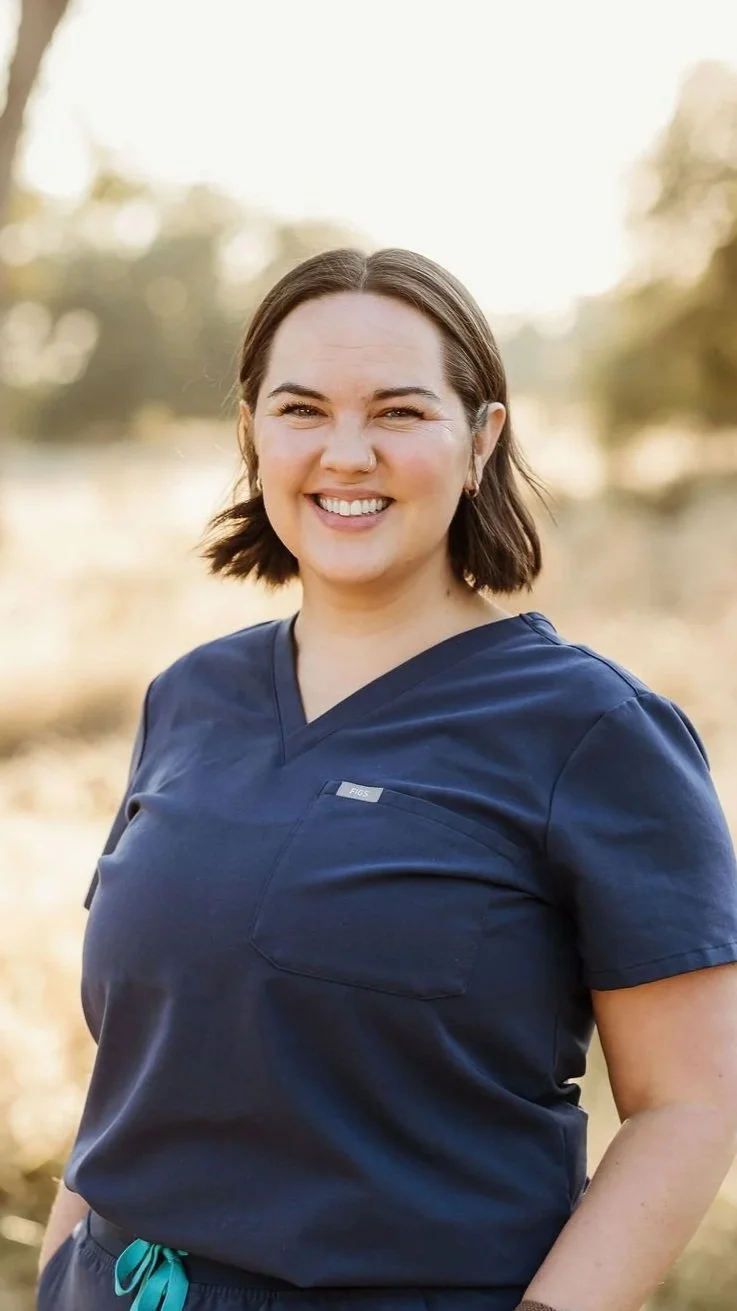 A smiling woman with shoulder-length brown hair, wearing blue scrubs, standing outdoors with trees and sunlight in the background.