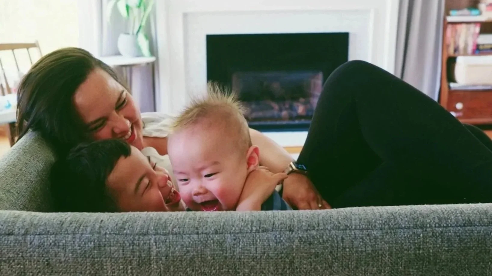 A woman and two young children playing and laughing on a gray couch in a living room with a fireplace, bookshelf, and window with a plant in the background.