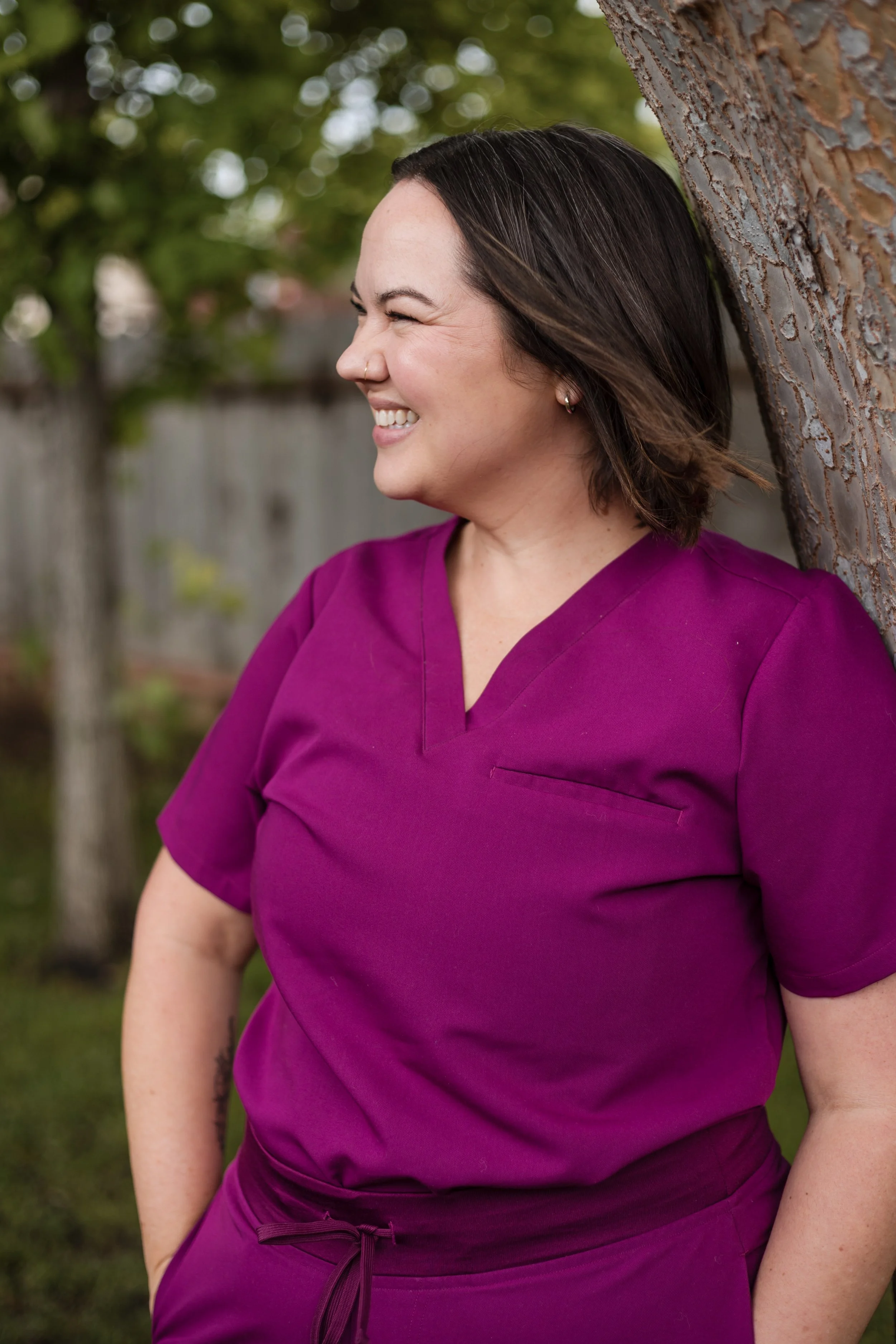 A woman with dark hair, wearing a purple medical scrub top, smiling and leaning against a tree outdoors.