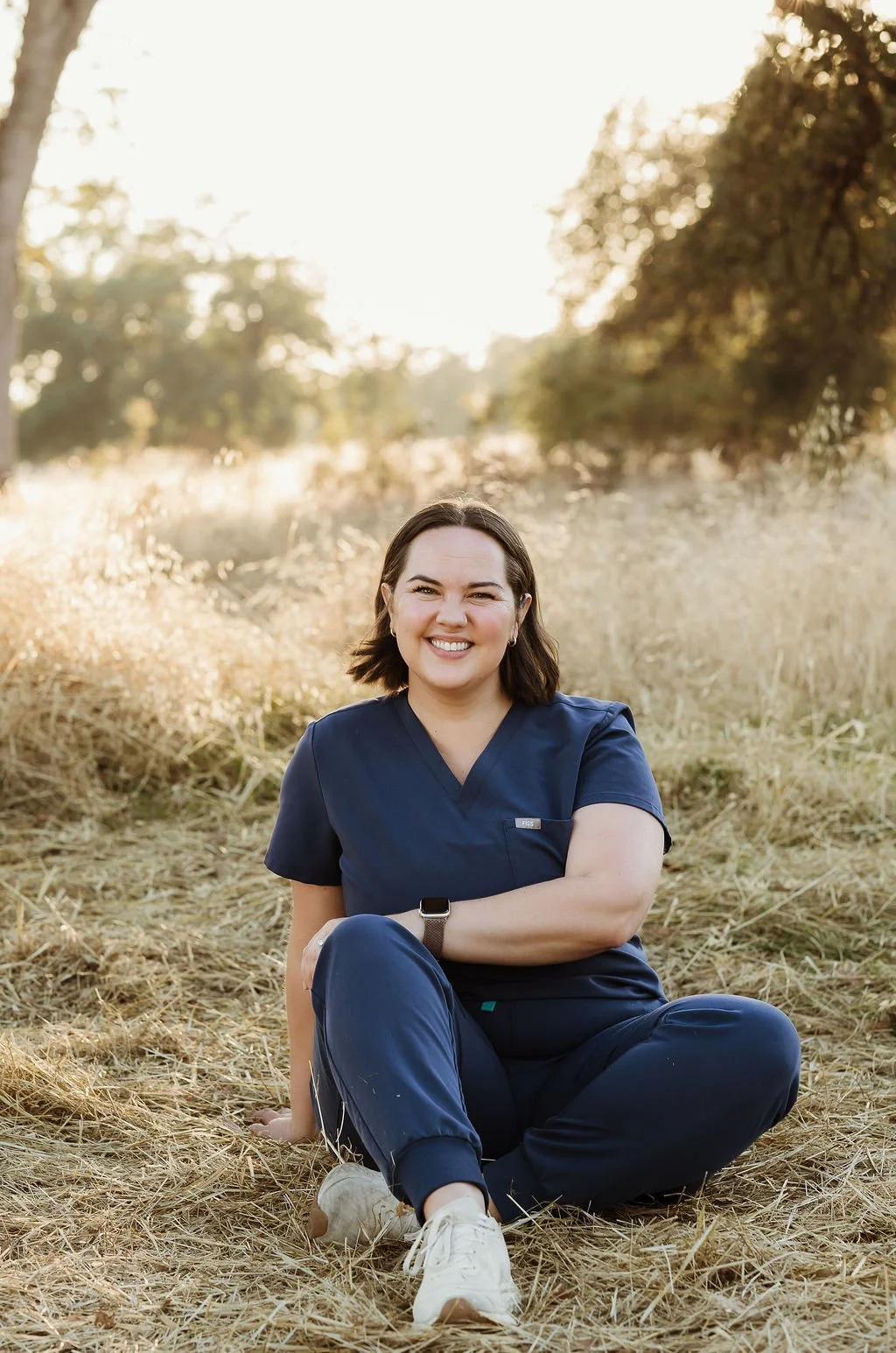 A woman in navy scrubs sitting on a grassy field at sunset, smiling and looking at the camera.