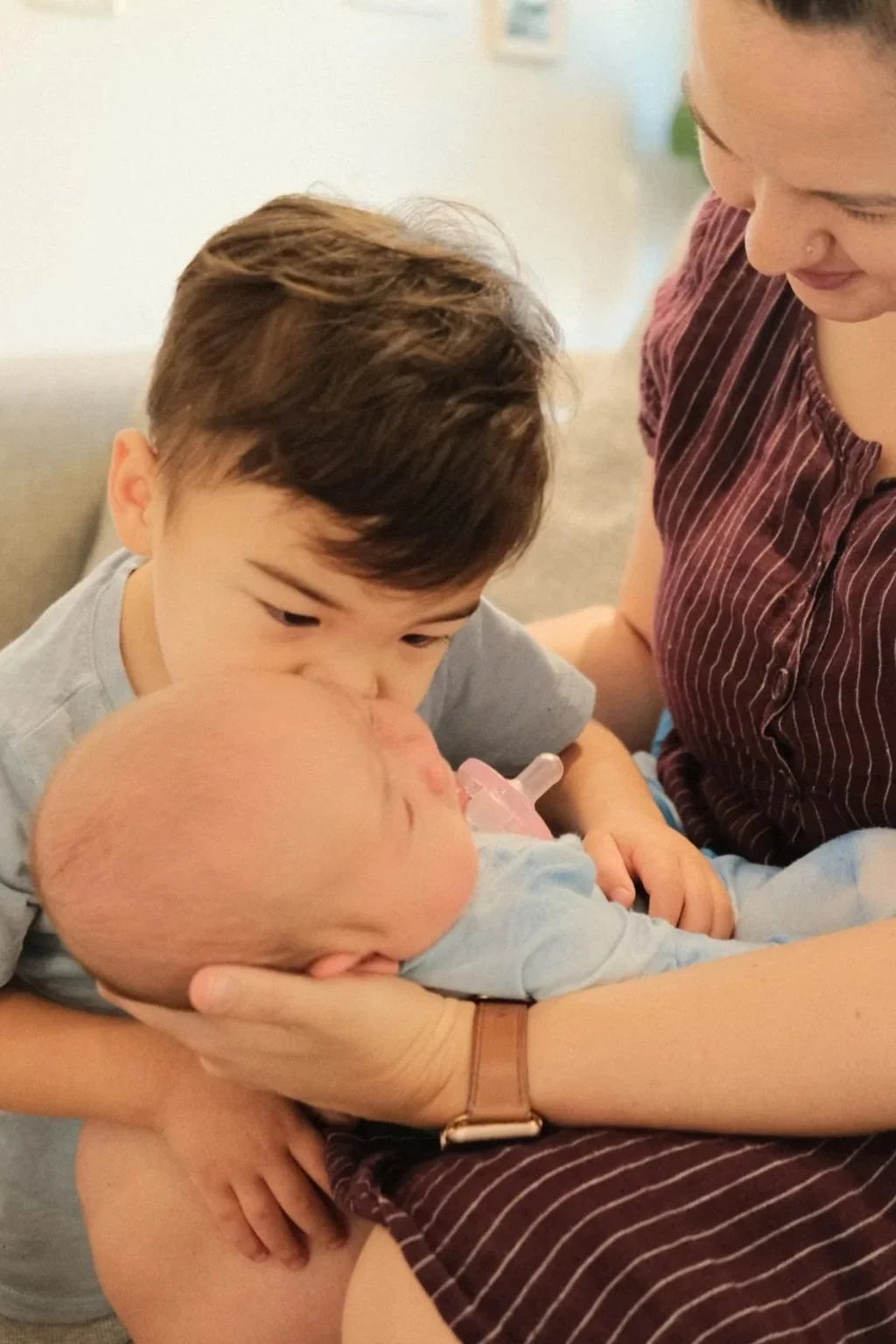 A young boy is holding an infant with a pacifier, while a woman looks on lovingly.