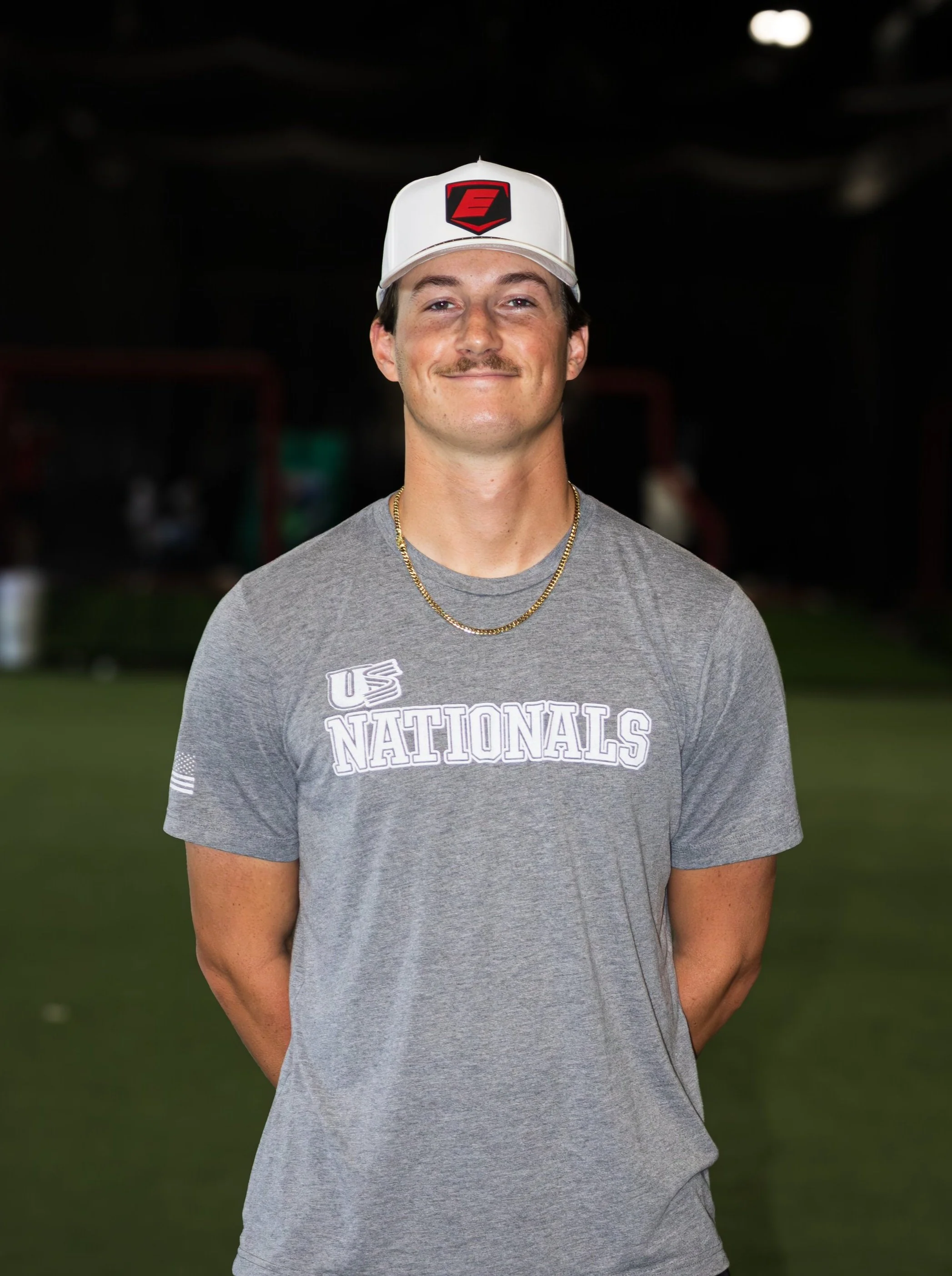 A young man wearing a white cap with a red and black logo, a gray T-shirt with 'US NATIONALS' text, and a gold necklace, standing outdoors at night.