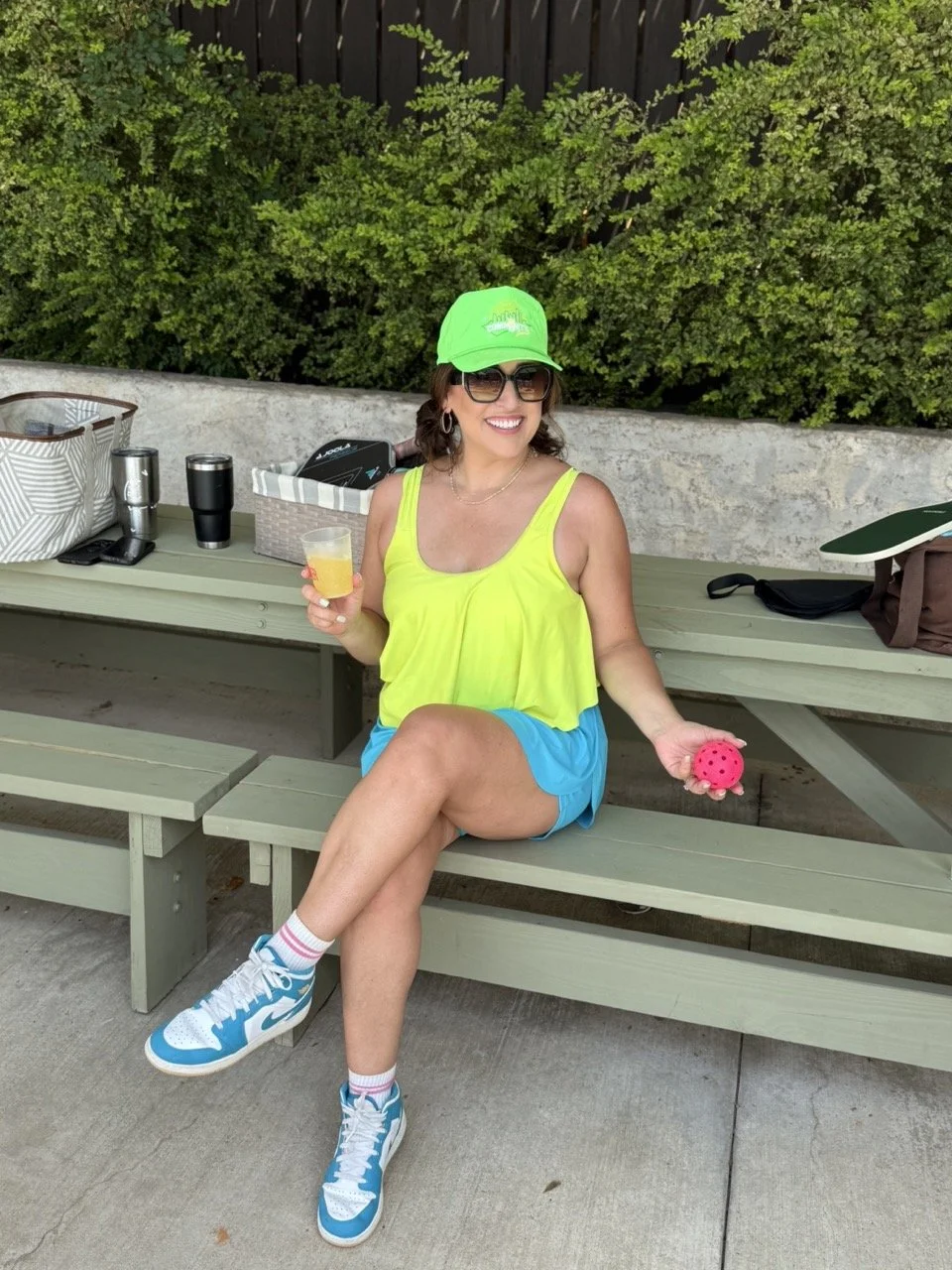 Woman sitting on a green wooden bench outdoors, smiling, wearing a lime green tank top, blue shorts, white and blue sneakers, a bright green cap, sunglasses, holding a drink and a pink ball.