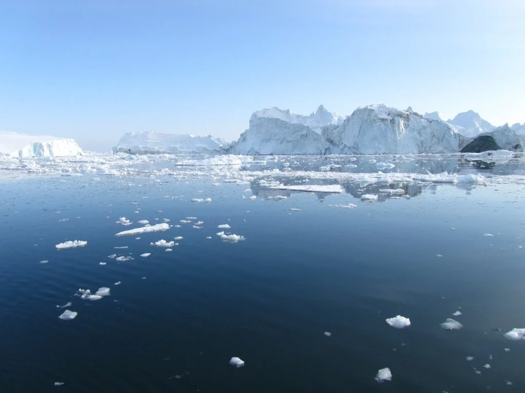 An icy seascape with large icebergs and floating ice chunks in calm blue water under a clear sky.