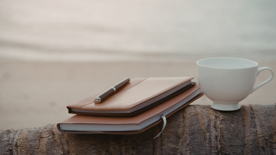 Notebook and pen beside a coffee cup with a calm lake in the background, suggesting reflection and writing