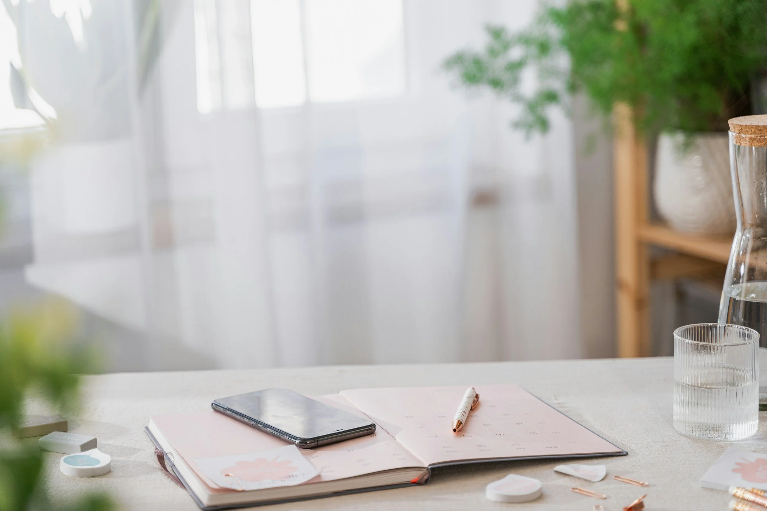 Open notebook on a desk with a pen, phone, and glass of water near a window.